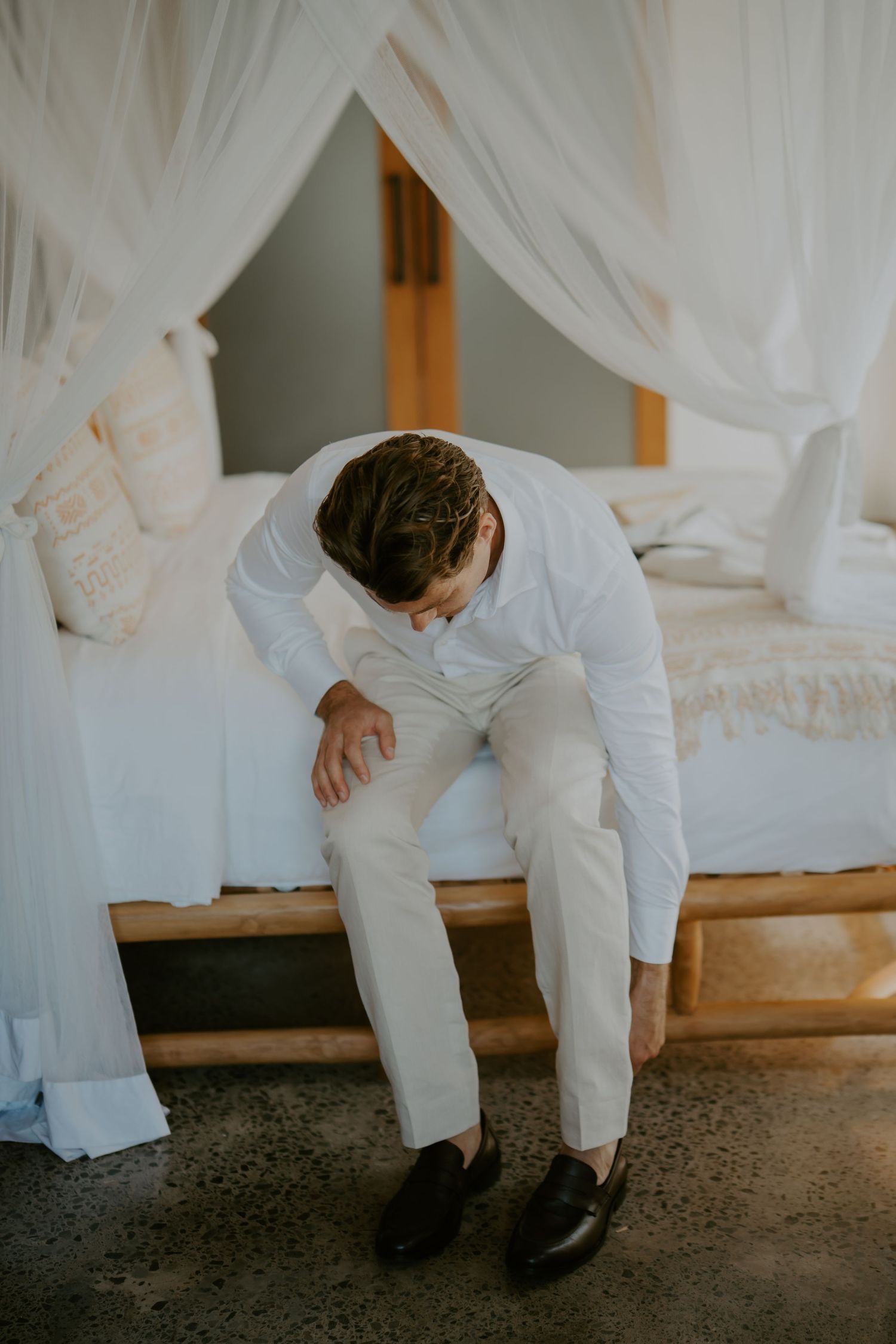 Groom in white linen suit sitting on a canopy bed adjusting his shoes before the wedding ceremony.