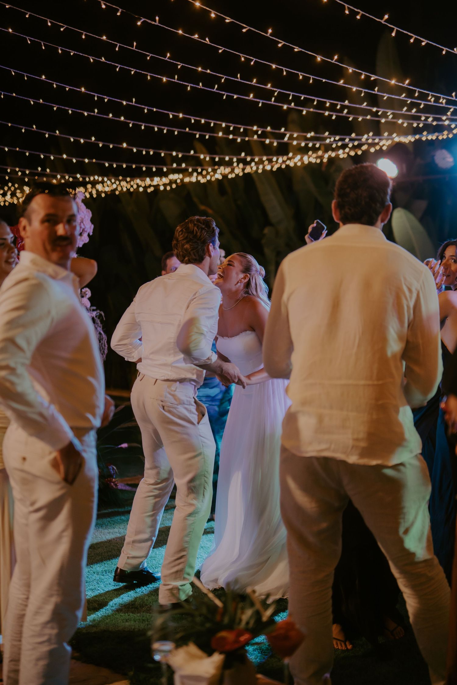Bride and groom share a joyful dance surrounded by guests in white attire under string lights at outdoor wedding reception.