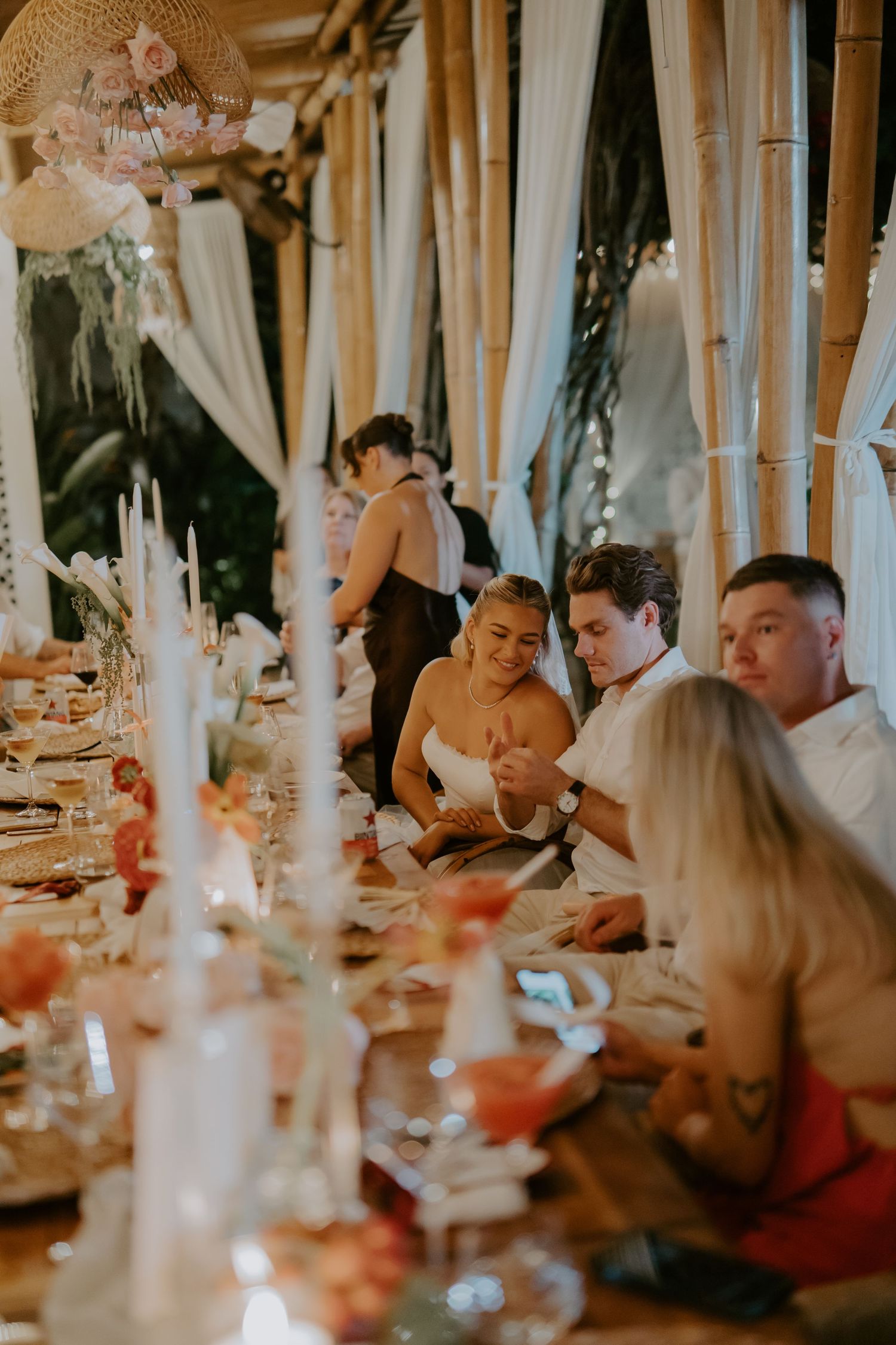 Wedding guests seated at a candlelit reception table decorated with flowers in a bamboo venue.