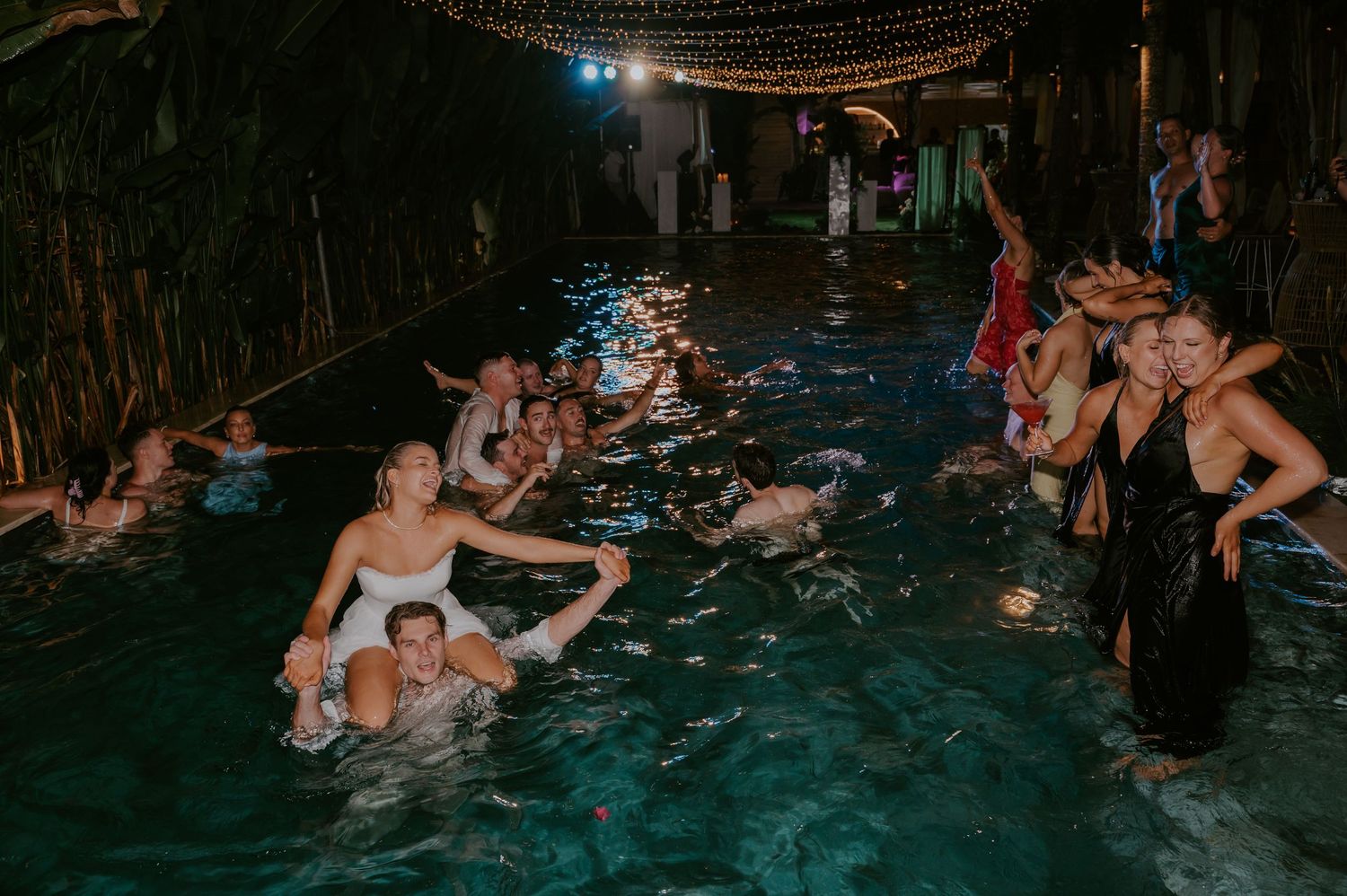 Wedding guests celebrate in illuminated pool at night during fun outdoor reception after-party.