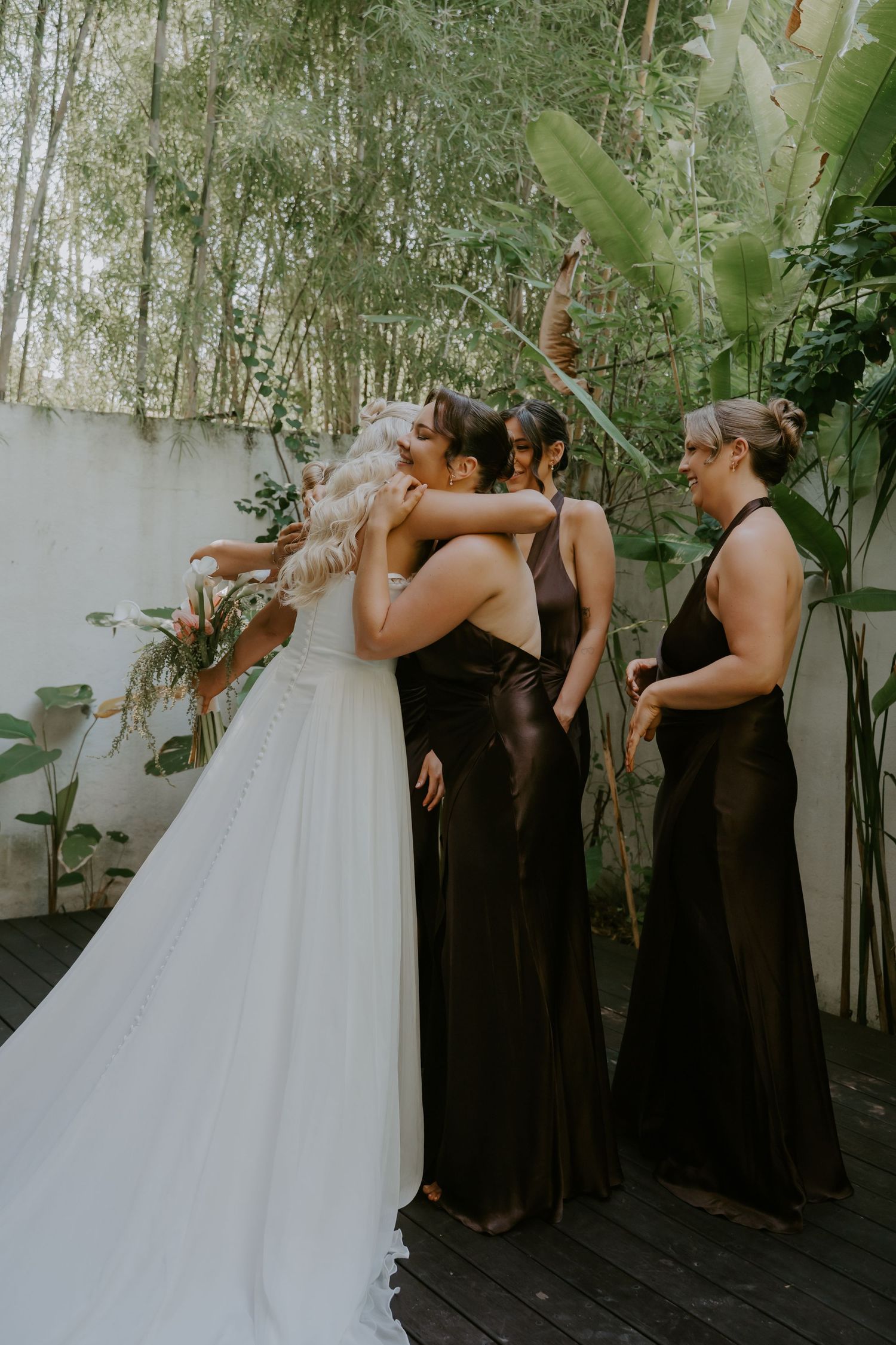 Bride in white gown embraces bridesmaids in dark dresses surrounded by tropical greenery.