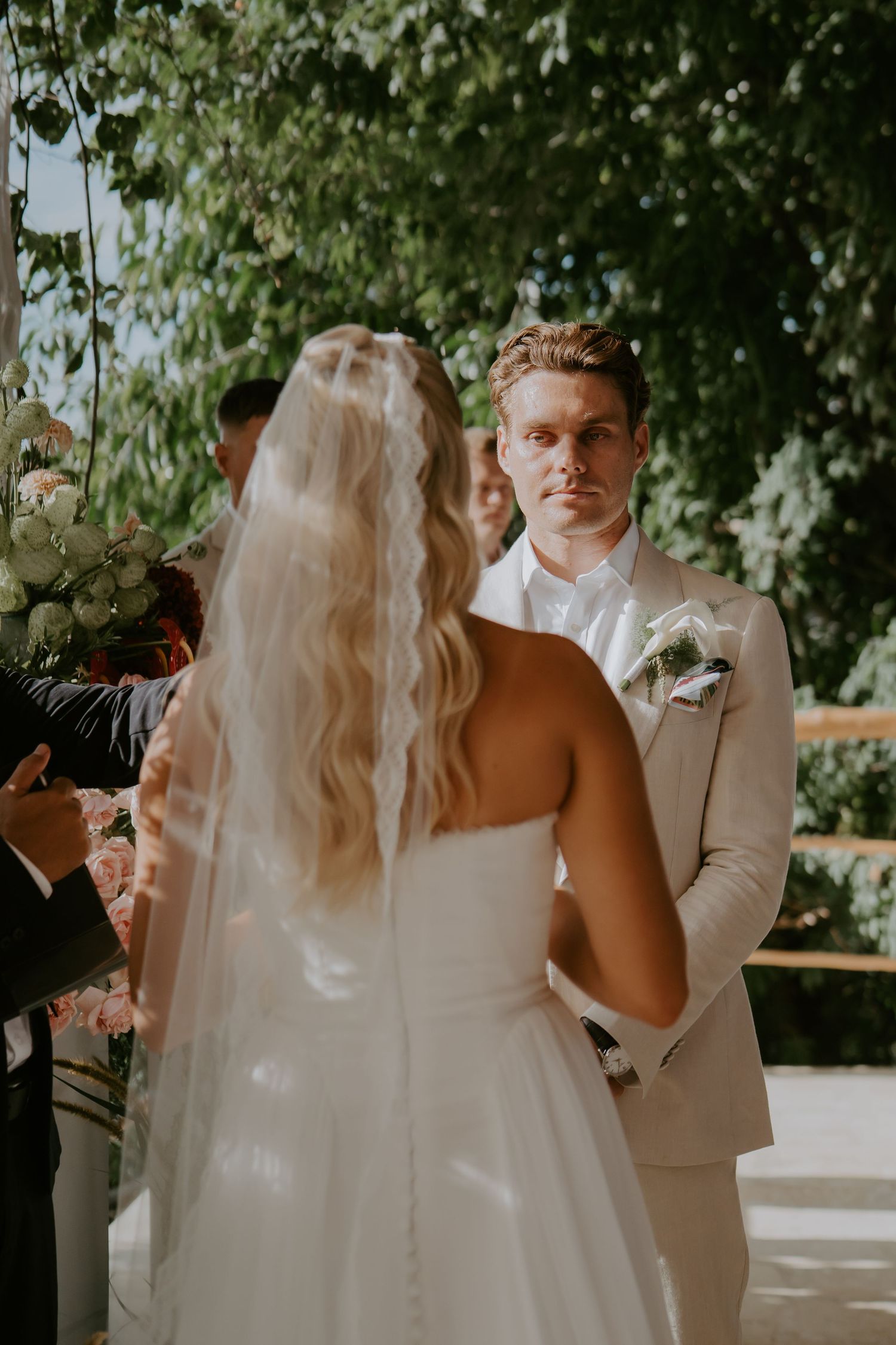 Groom in beige suit gazes at bride during outdoor wedding ceremony surrounded by lush greenery.