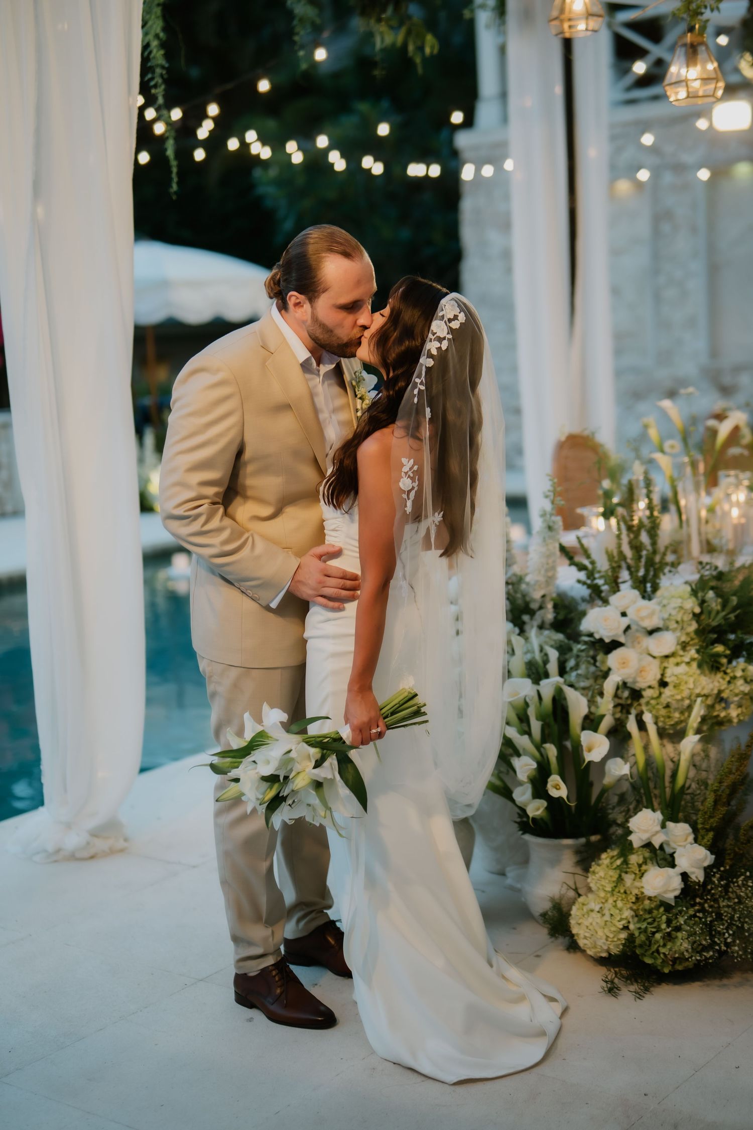 Bride and groom sharing a kiss at elegant outdoor wedding reception near pool with white floral arrangements.
