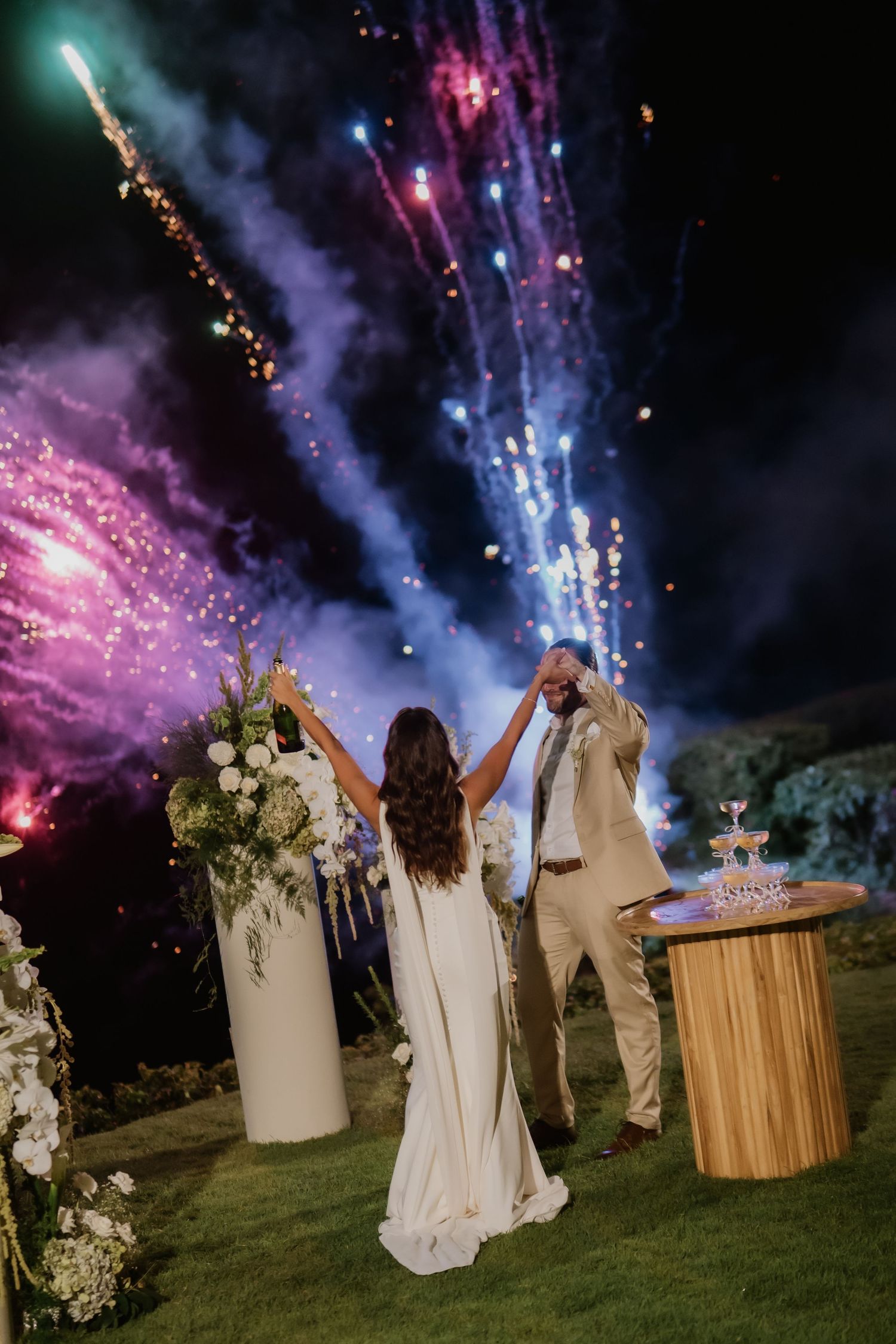Bride and groom celebrating with raised glasses during colorful fireworks display at night.