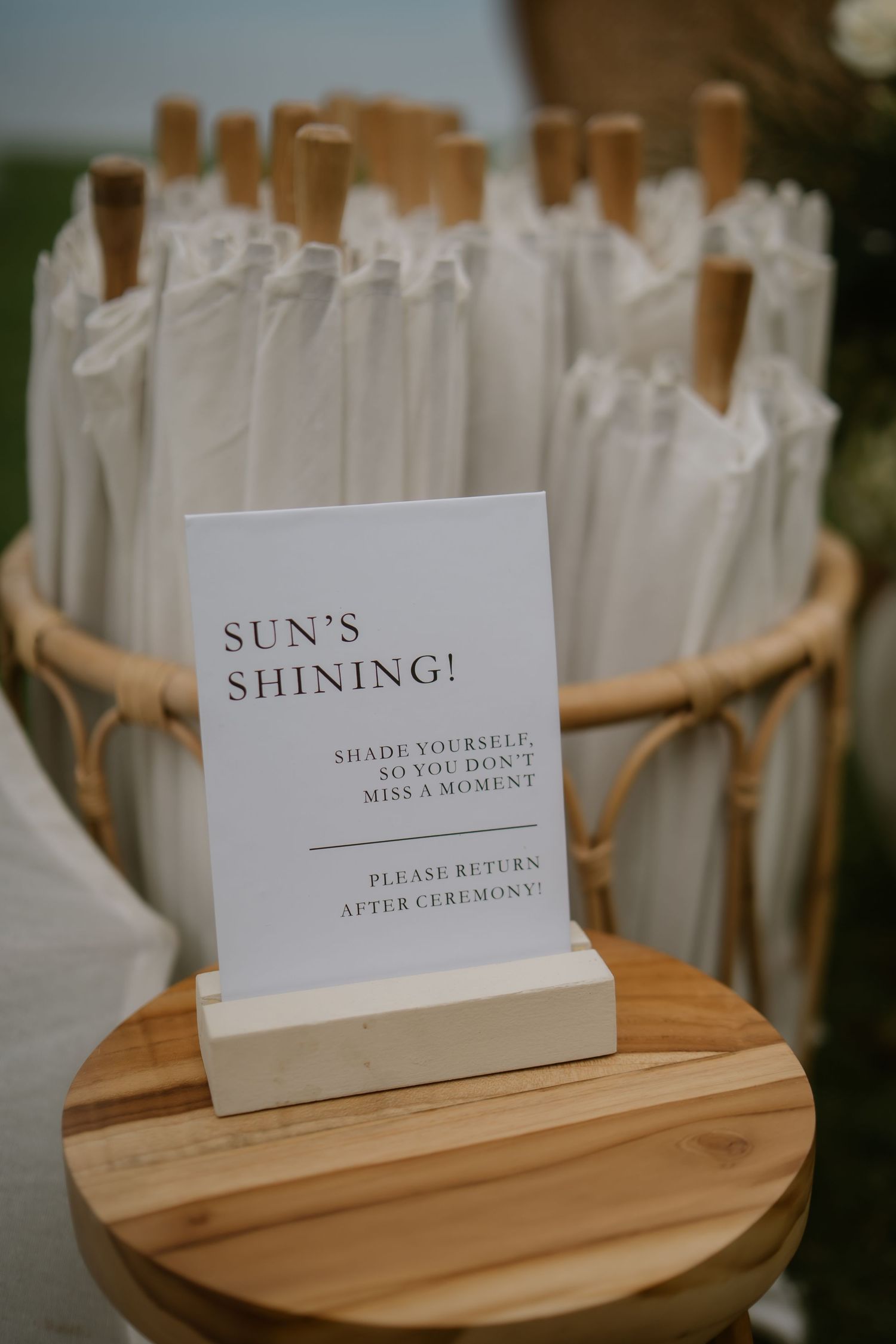 White parasols in a rattan basket with a sign reading Sun's Shining, shade yourself, please return after ceremony.