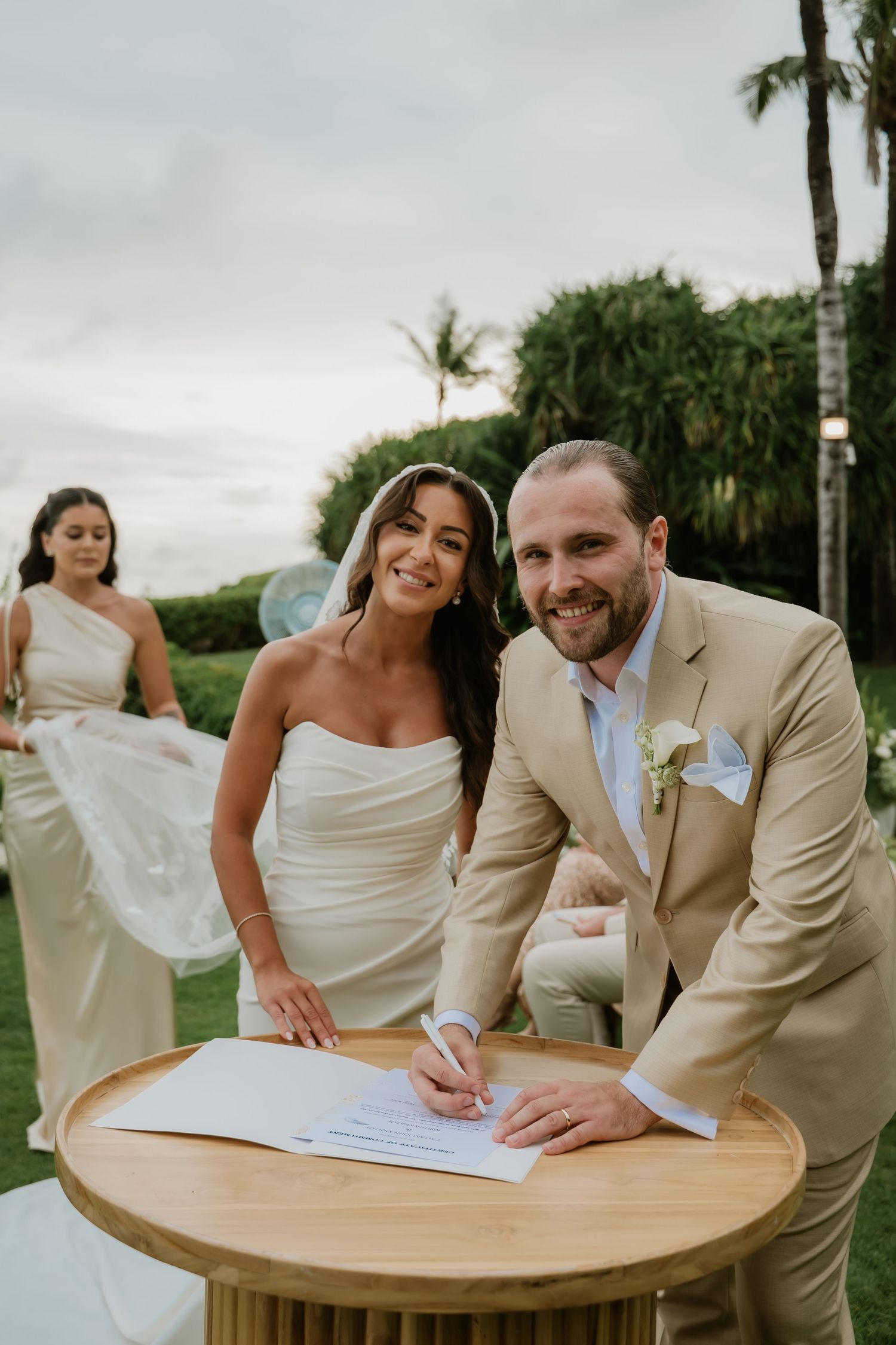 Bride and groom smiling while signing wedding certificate at outdoor ceremony with tropical greenery backdrop.