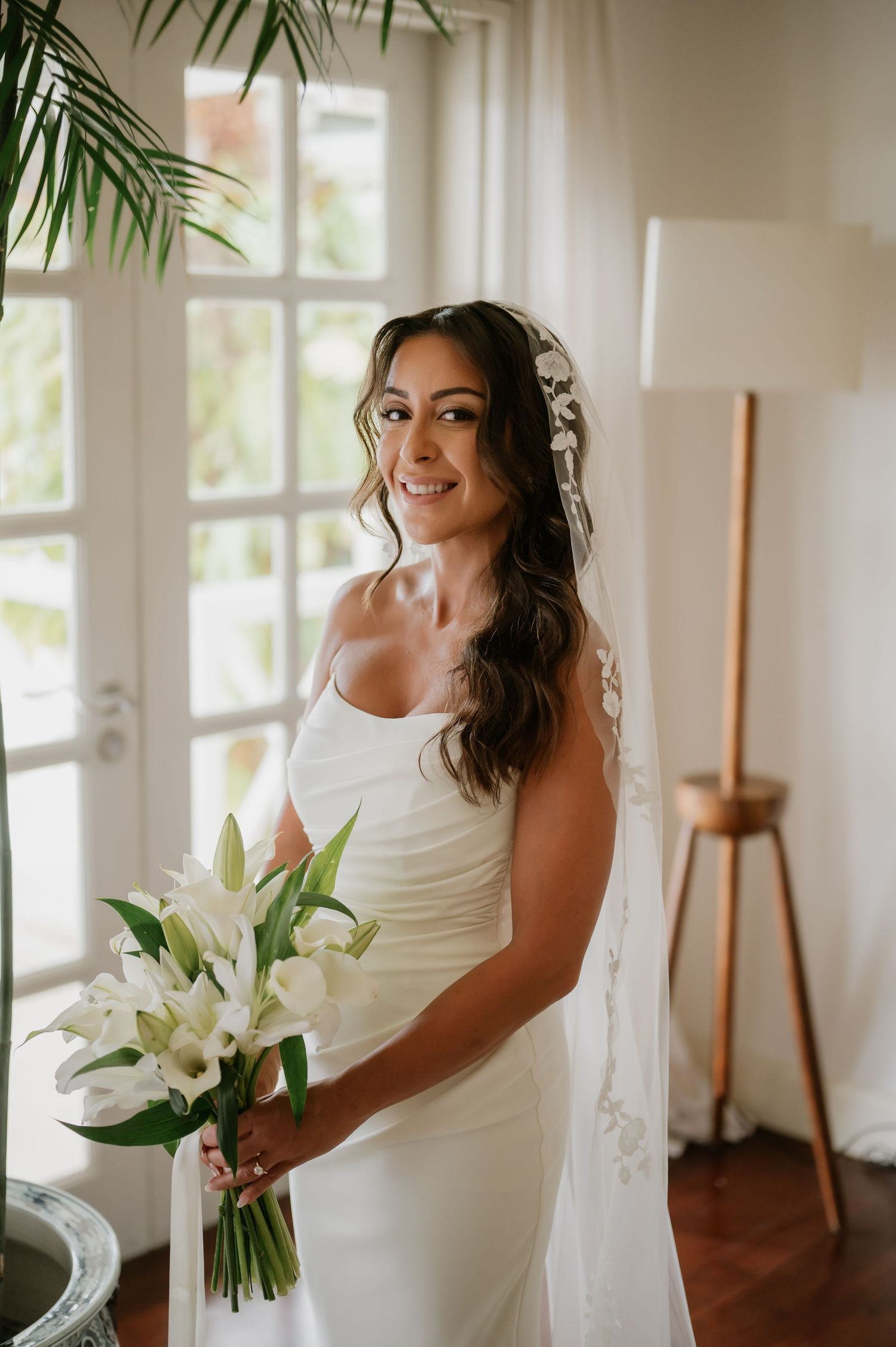 Smiling bride in a white gown holding white lily bouquet with lace veil on wedding day.