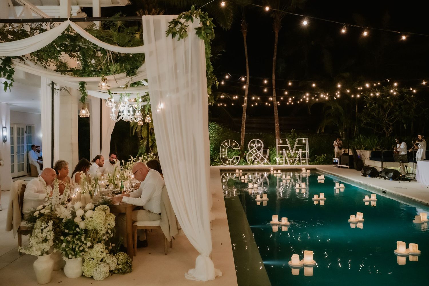 Outdoor wedding reception by pool at night with string lights and floral arrangements.