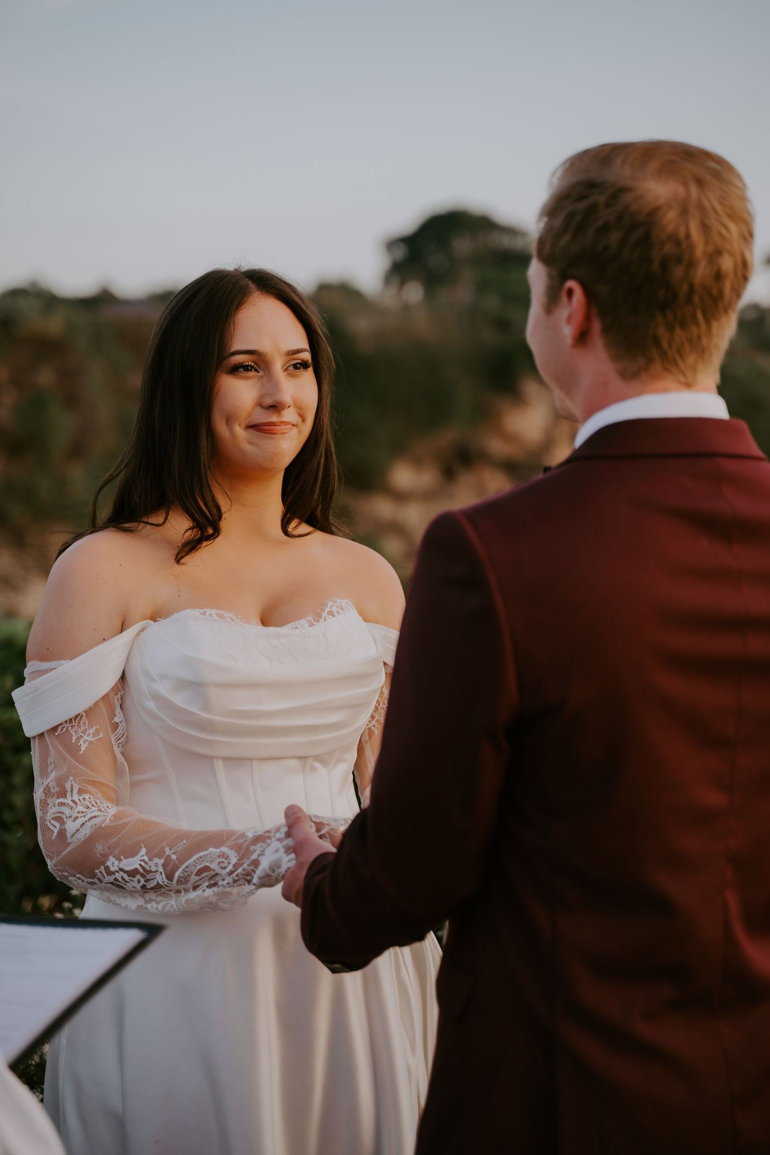 Smiling bride in off-shoulder lace gown holds grooms hands during ceremony.