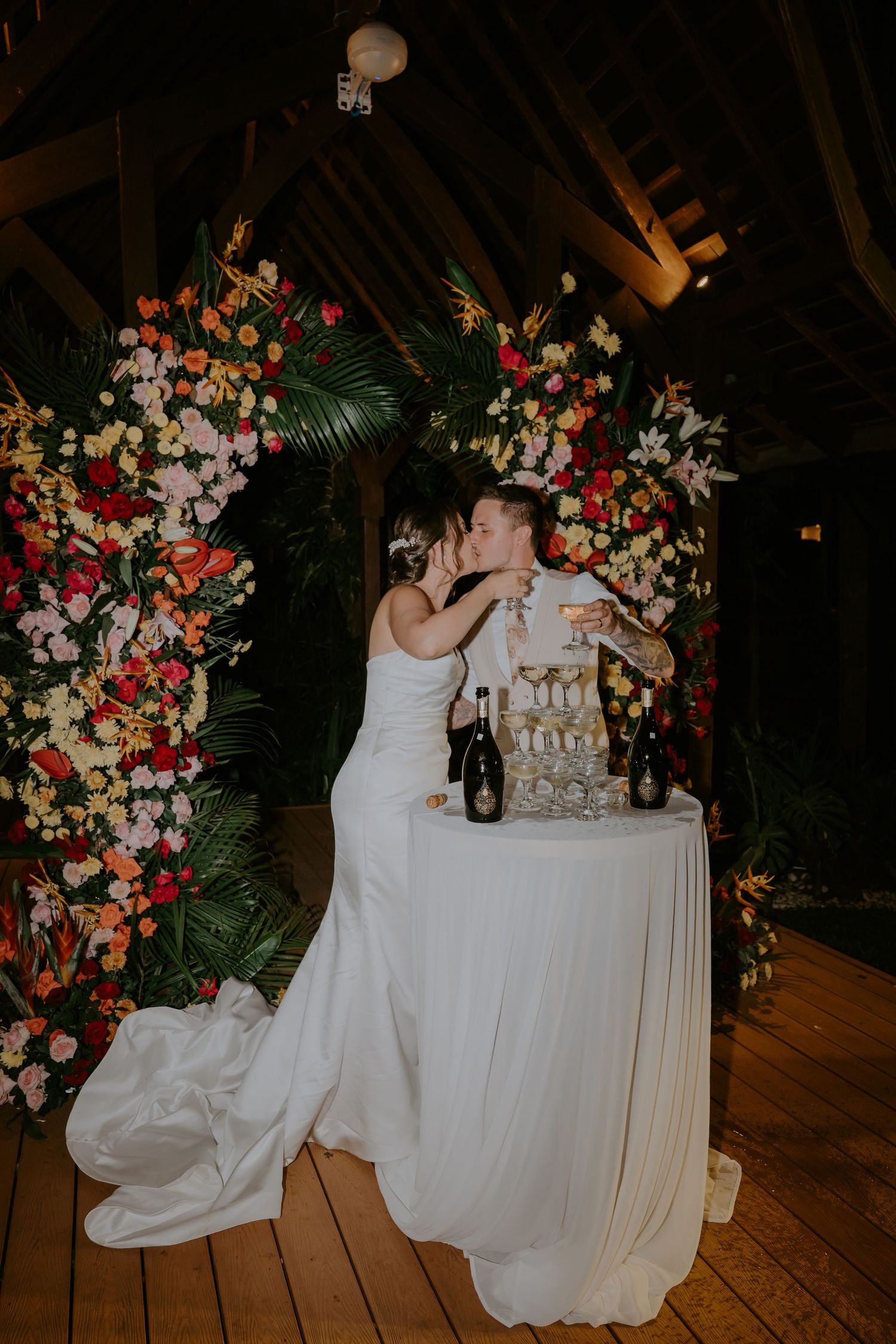 Bride and groom kissing beside champagne tower under colorful floral arch at wedding reception.