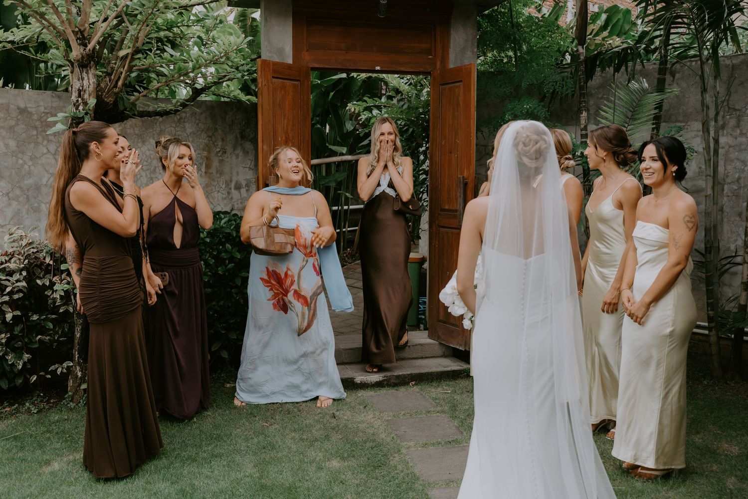 Bridesmaids in brown gowns react with joy as bride in white dress and veil approaches.