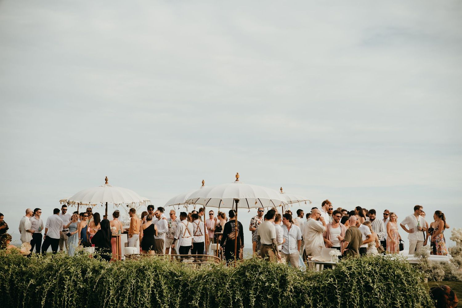 Wedding guests in white attire mingling outdoors under decorative tent canopy at seaside venue.