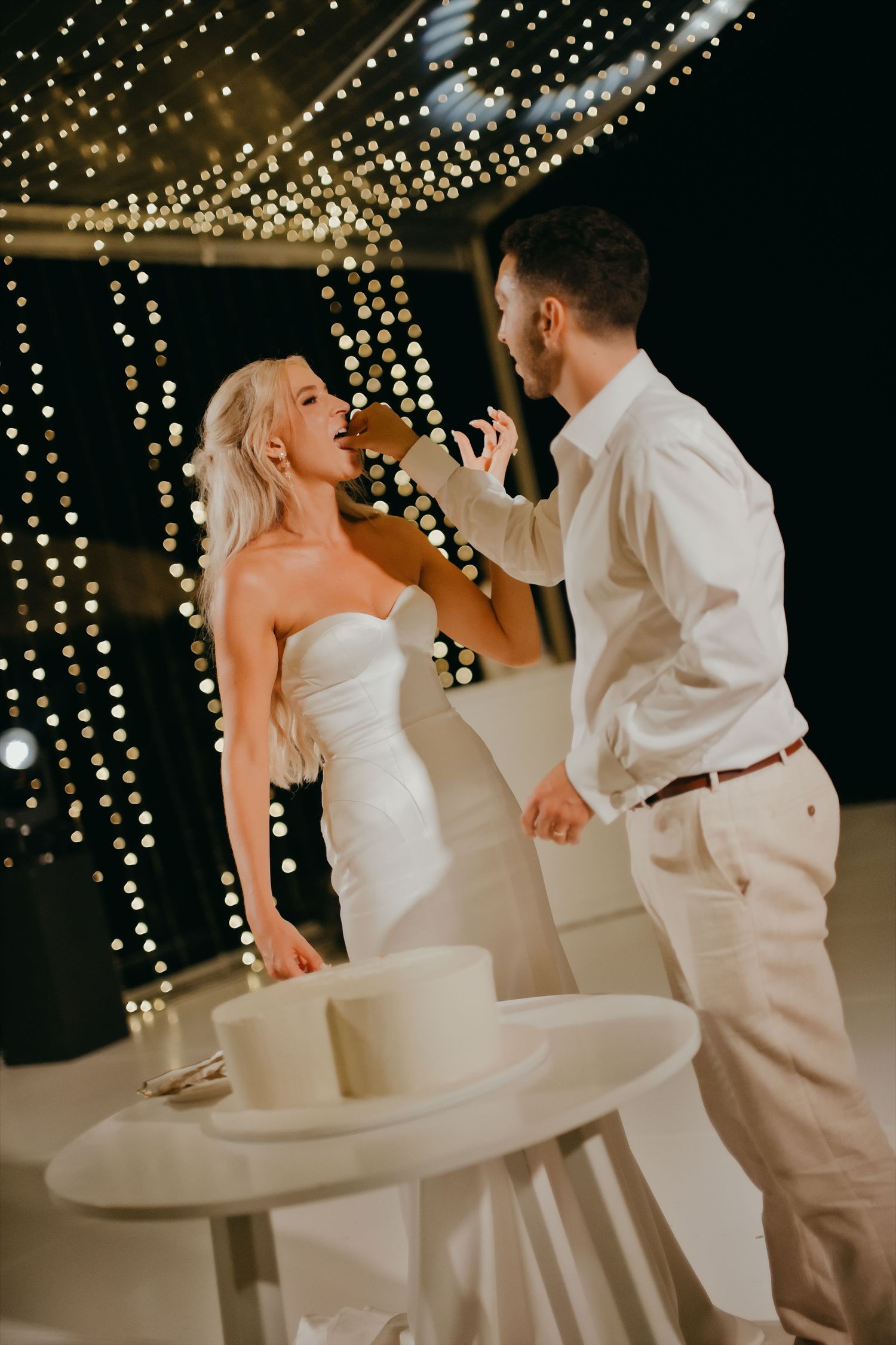 Groom feeding bride a piece of wedding cake under a canopy of sparkling fairy lights at night.