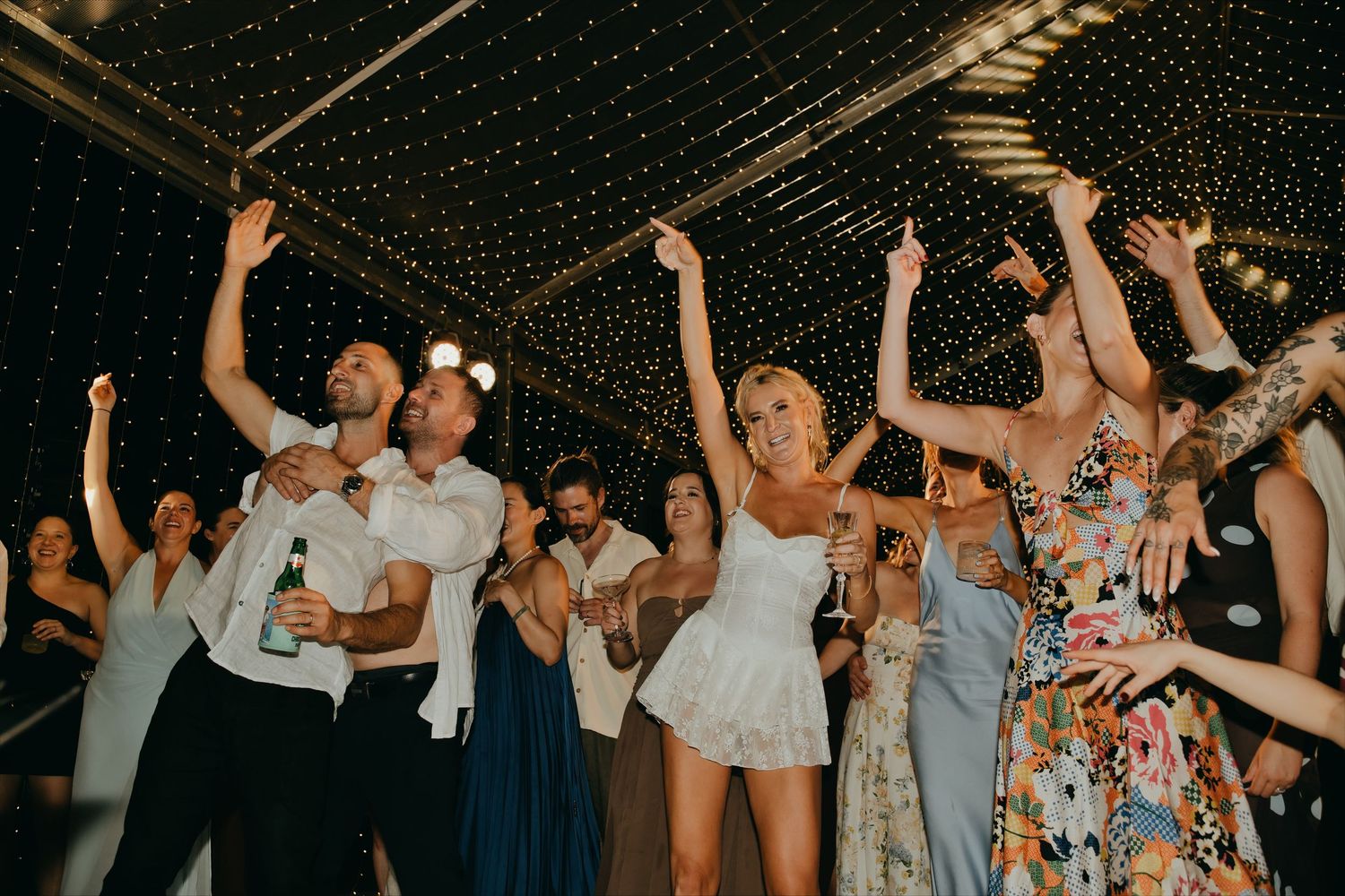 Wedding guests dance joyfully with hands raised under starlit ceiling at lively indoor reception party.