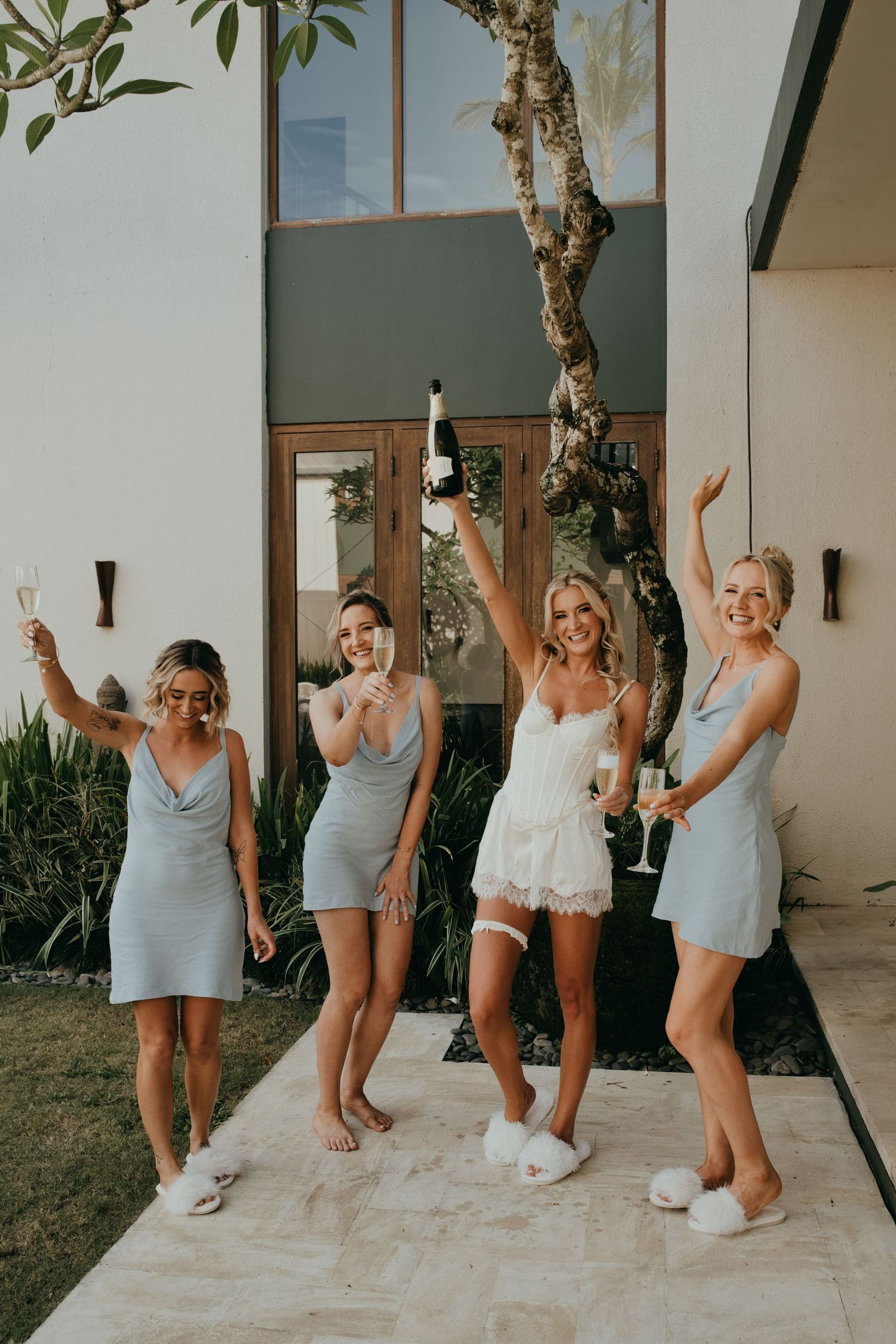 Bride in white lace romper celebrates with bridesmaids in blue satin dresses holding champagne glasses.