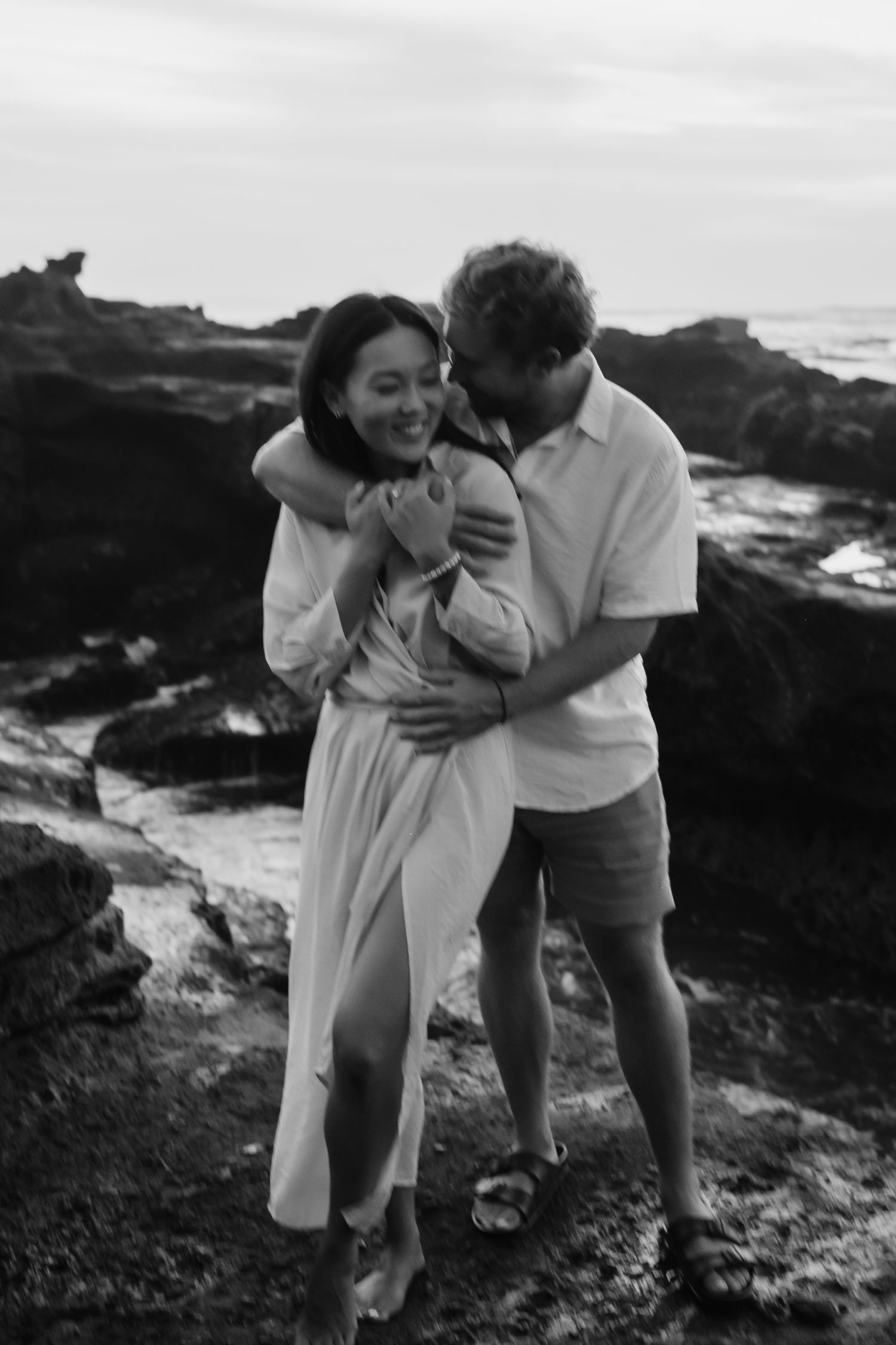 A couple shares a romantic moment on coastal rocks in black and white photography.