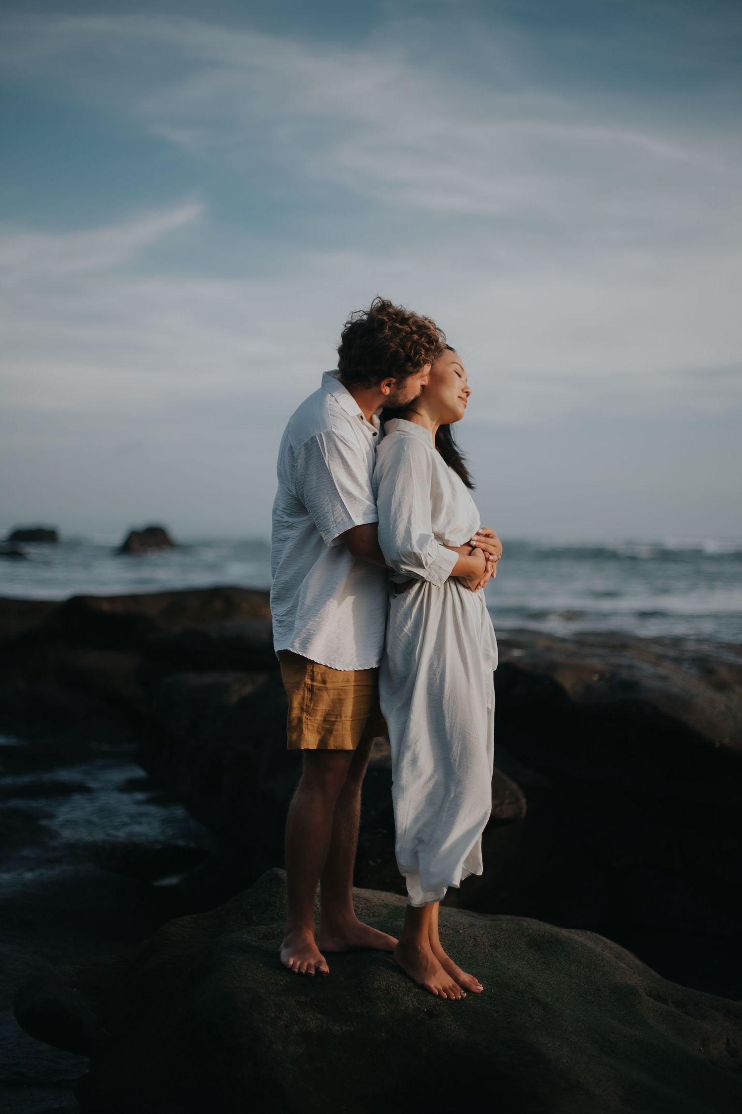 Romantic couple embraces on rocky beach at sunset wearing light colored clothing with ocean waves in background.