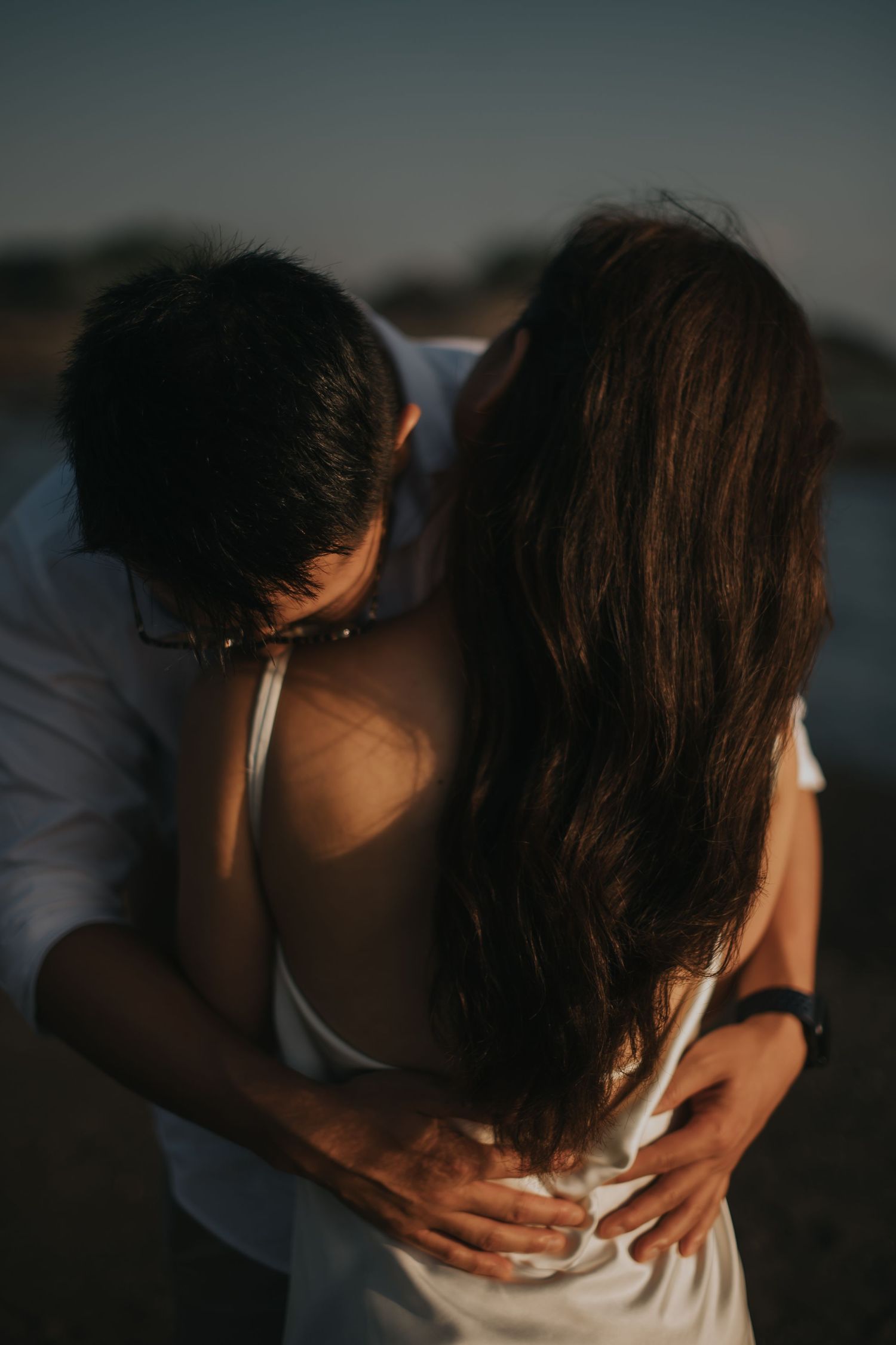 Romantic couple embracing at sunset, man in white shirt kissing woman with long dark hair near water.