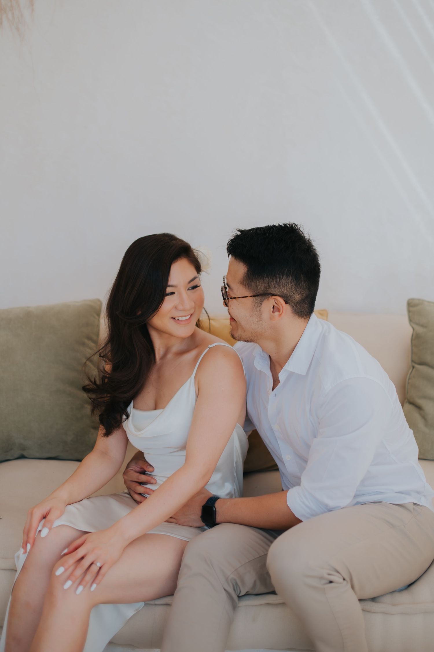 A smiling Asian couple sits on a cozy sofa, the man whispering to the woman in a white dress during a romantic photo session.