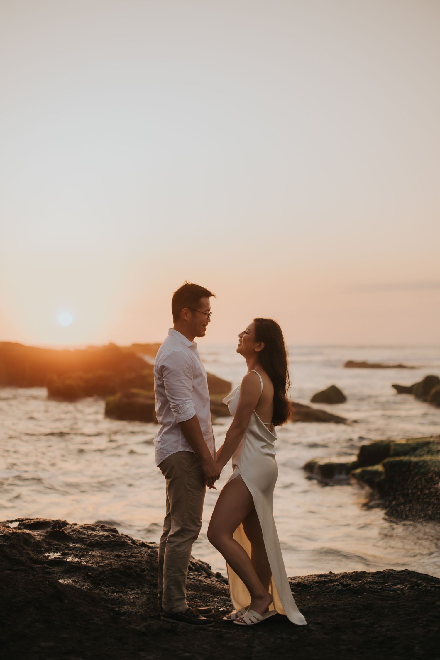 Couple holding hands on rocky beach at golden sunset, woman in white dress and man in white shirt facing each other romantically.