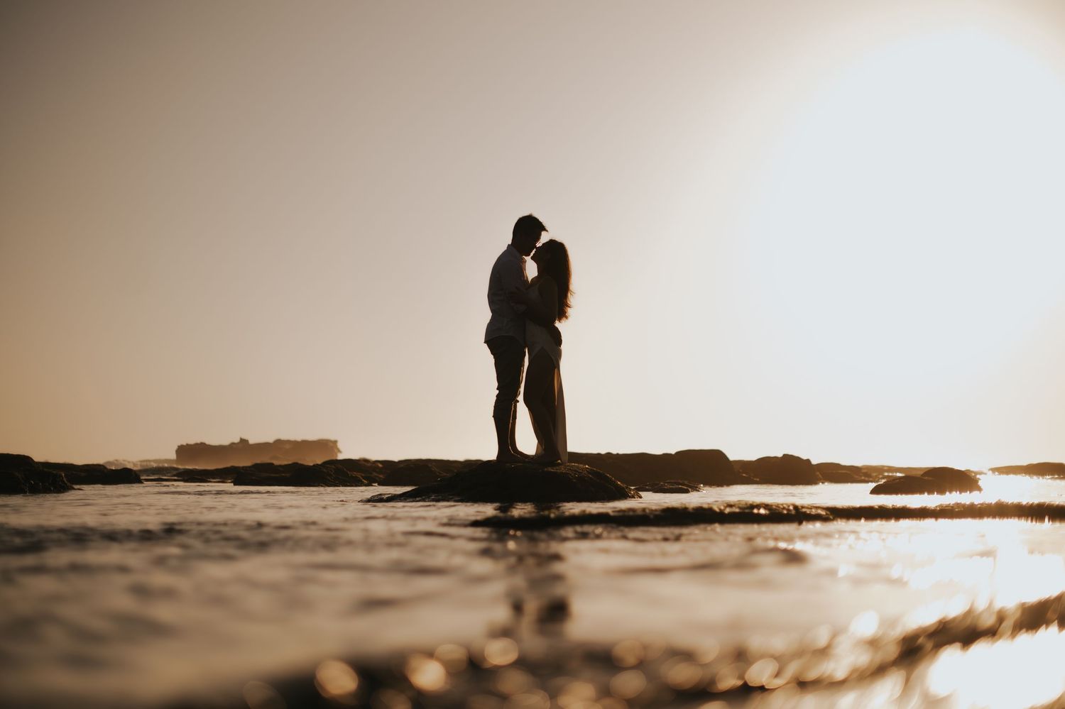 Silhouette of a couple kissing on rocky shore at golden sunset with ocean waves surrounding them.