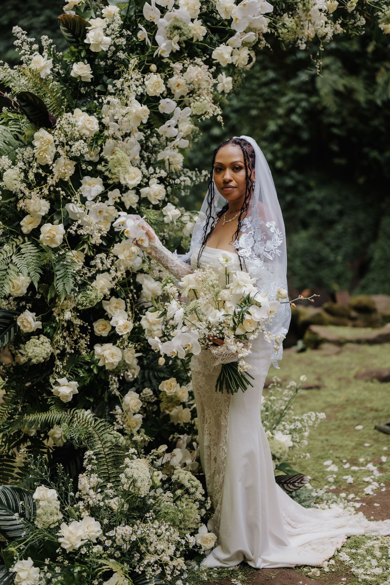 Elegant bride in lace wedding dress and veil holds white bouquet beside a stunning white floral arch outdoors.