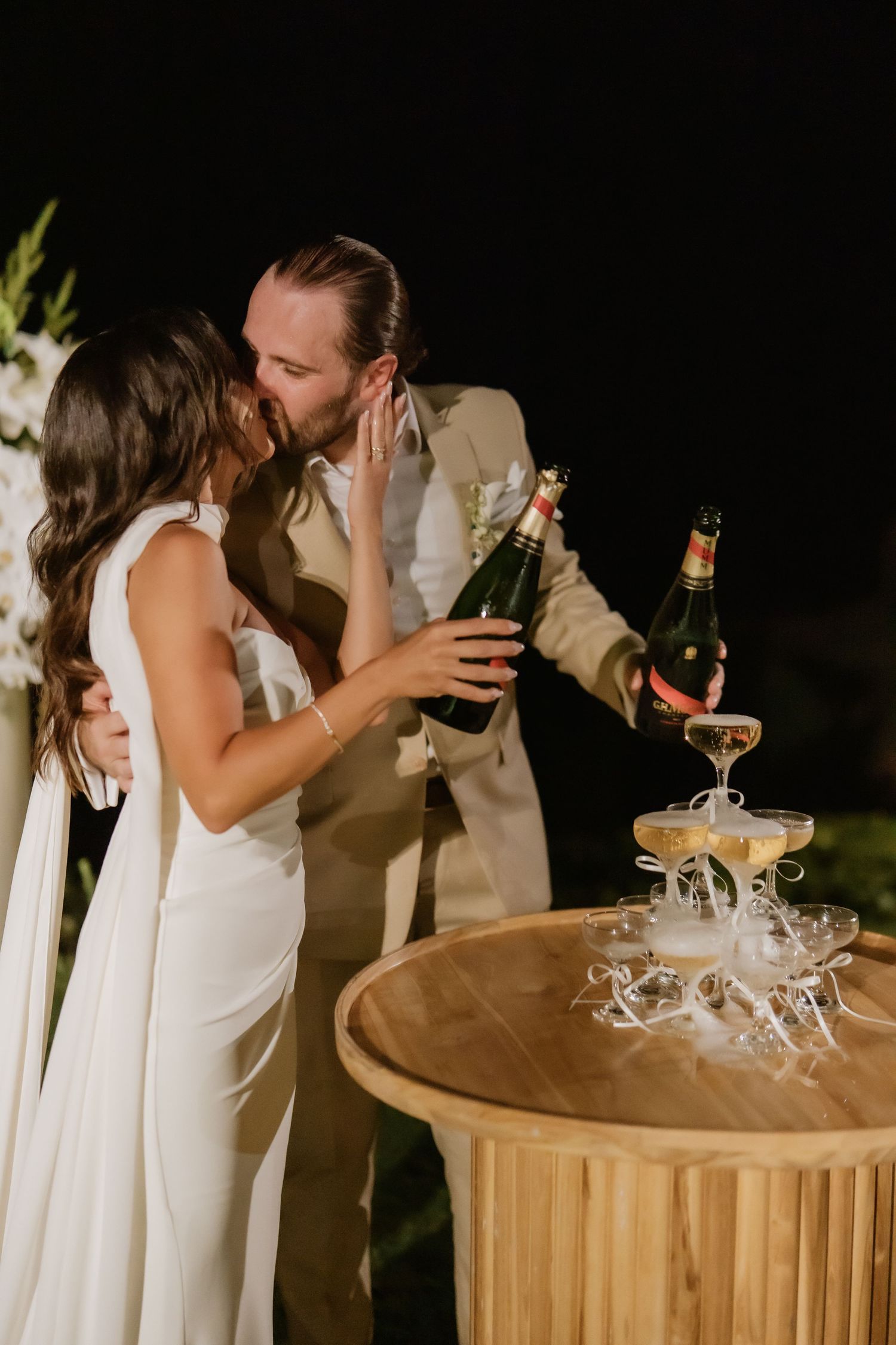 Newlyweds kiss while pouring champagne tower at outdoor night wedding reception.