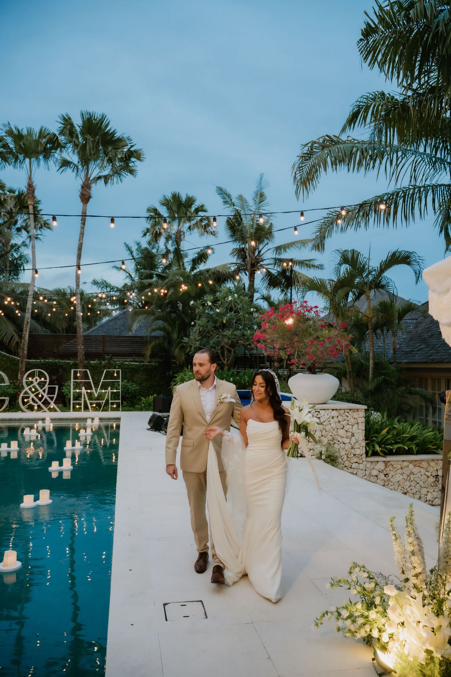 Bride and groom walk poolside at dusk under string lights surrounded by tropical palm trees.