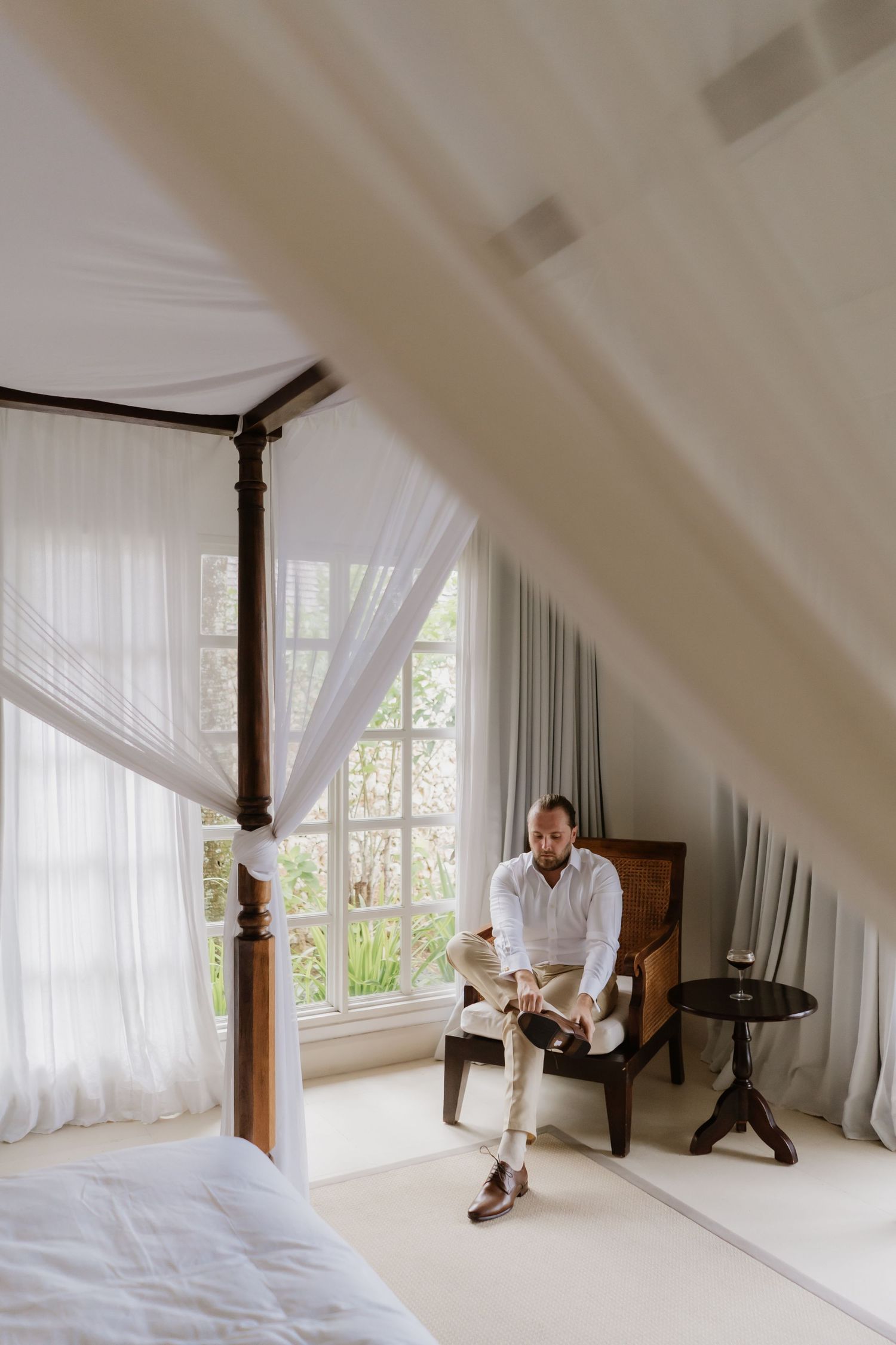 A groom in a white shirt sits in a canopy bedroom tying his shoes before the wedding.