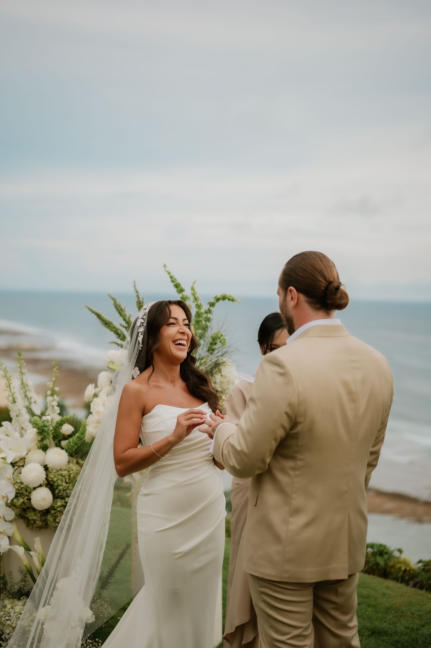 Bride laughing joyfully with groom during oceanfront wedding ceremony with floral arch backdrop.