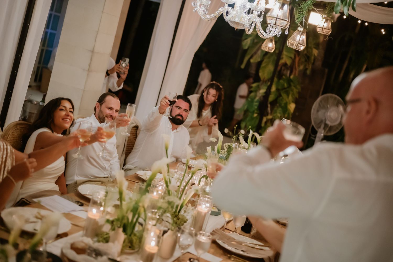 Wedding guests raising glasses in a joyful champagne toast at a candlelit reception dinner table.