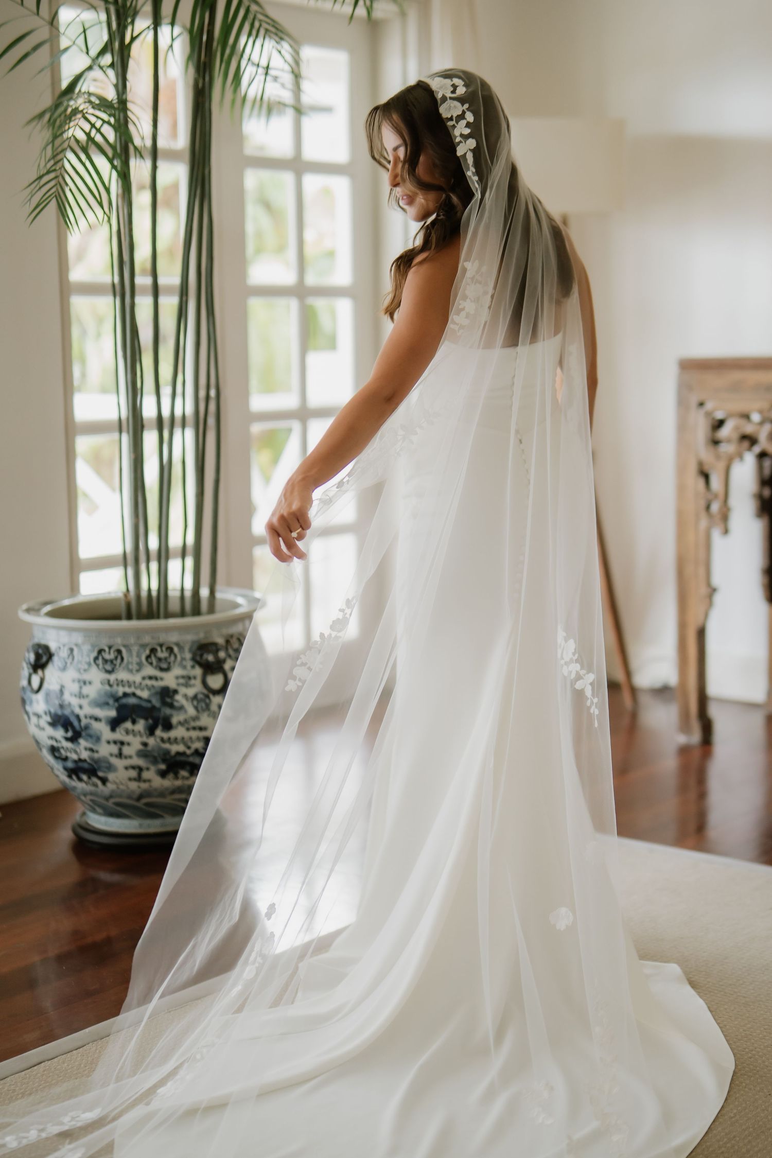 Bride in elegant white wedding gown with long floral veil posing in a bright sunlit room.