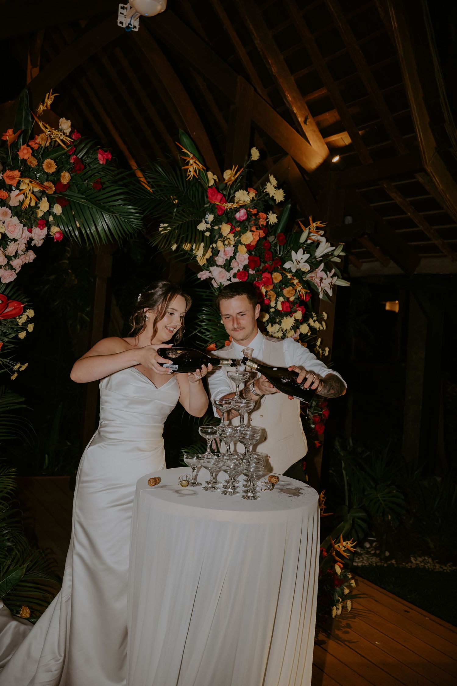 Bride and groom pour champagne tower at a tropical floral wedding reception indoors at night.