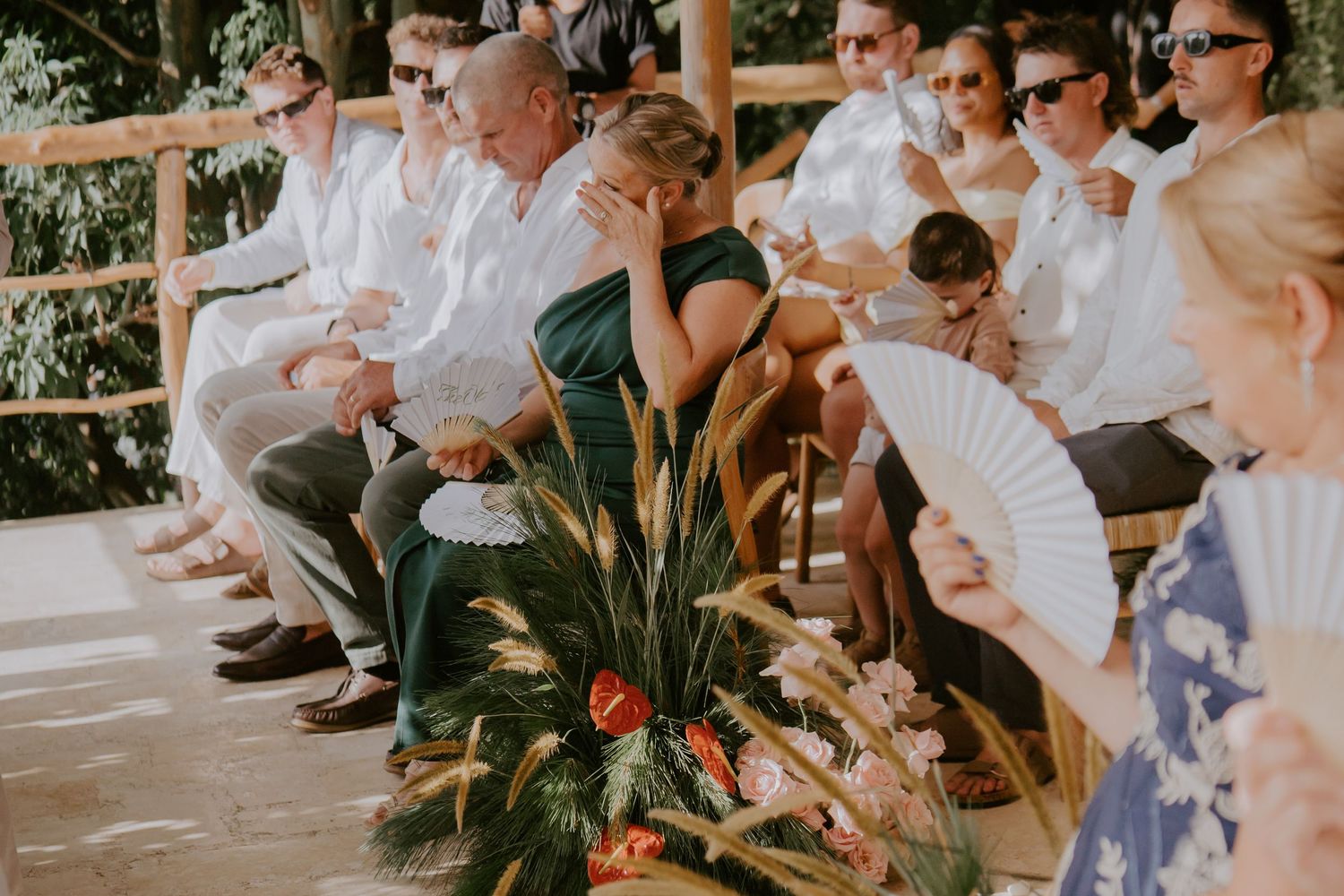 Wedding guests seated outdoors fanning themselves during tropical ceremony with floral arrangements in foreground.