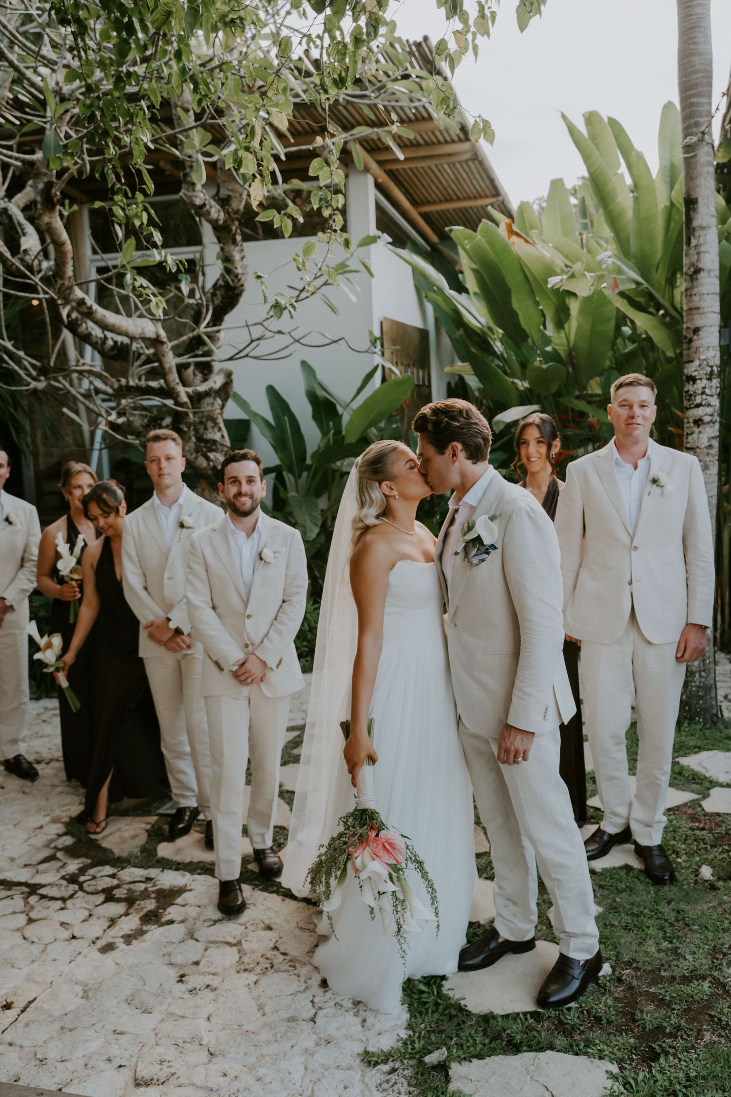 Bride and groom share first kiss surrounded by groomsmen in beige suits at outdoor ceremony.