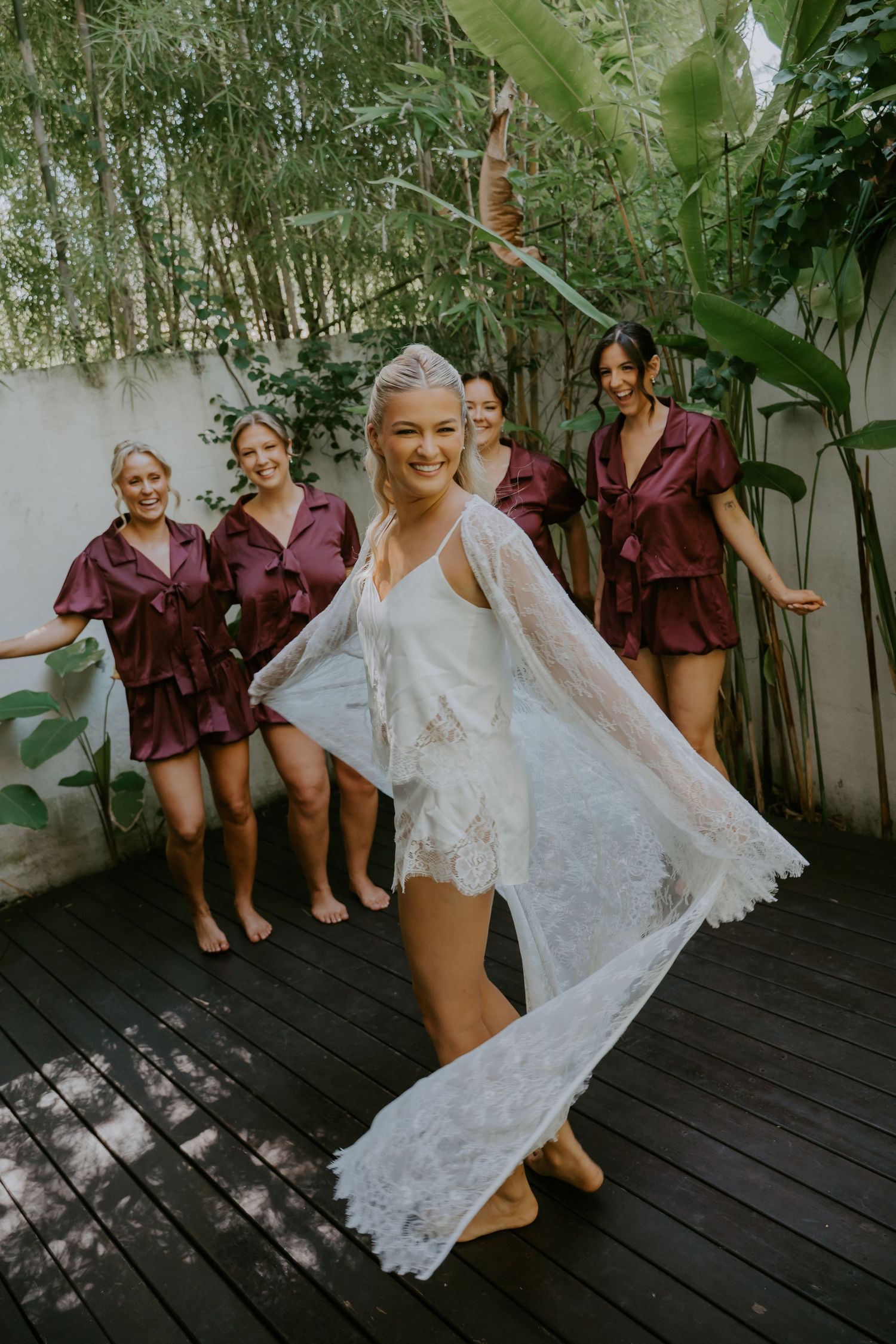 Bride in white lace robe holding veil with bridesmaids in burgundy satin robes on tropical outdoor deck.