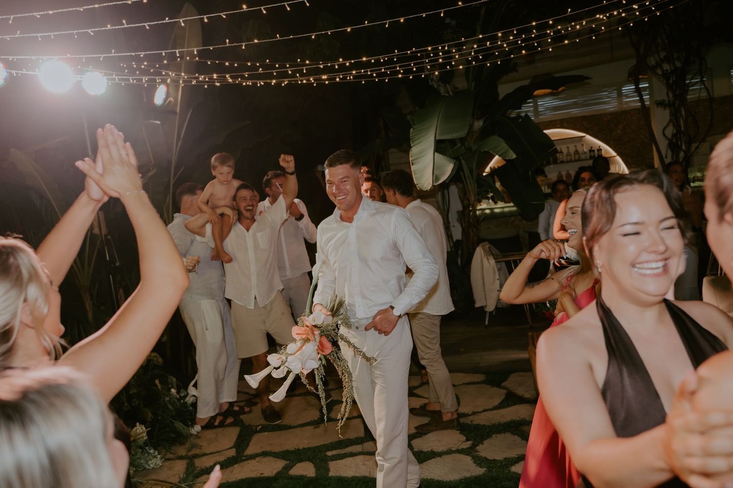 Groom in white suit celebrating joyfully with wedding guests during lively outdoor evening reception party.