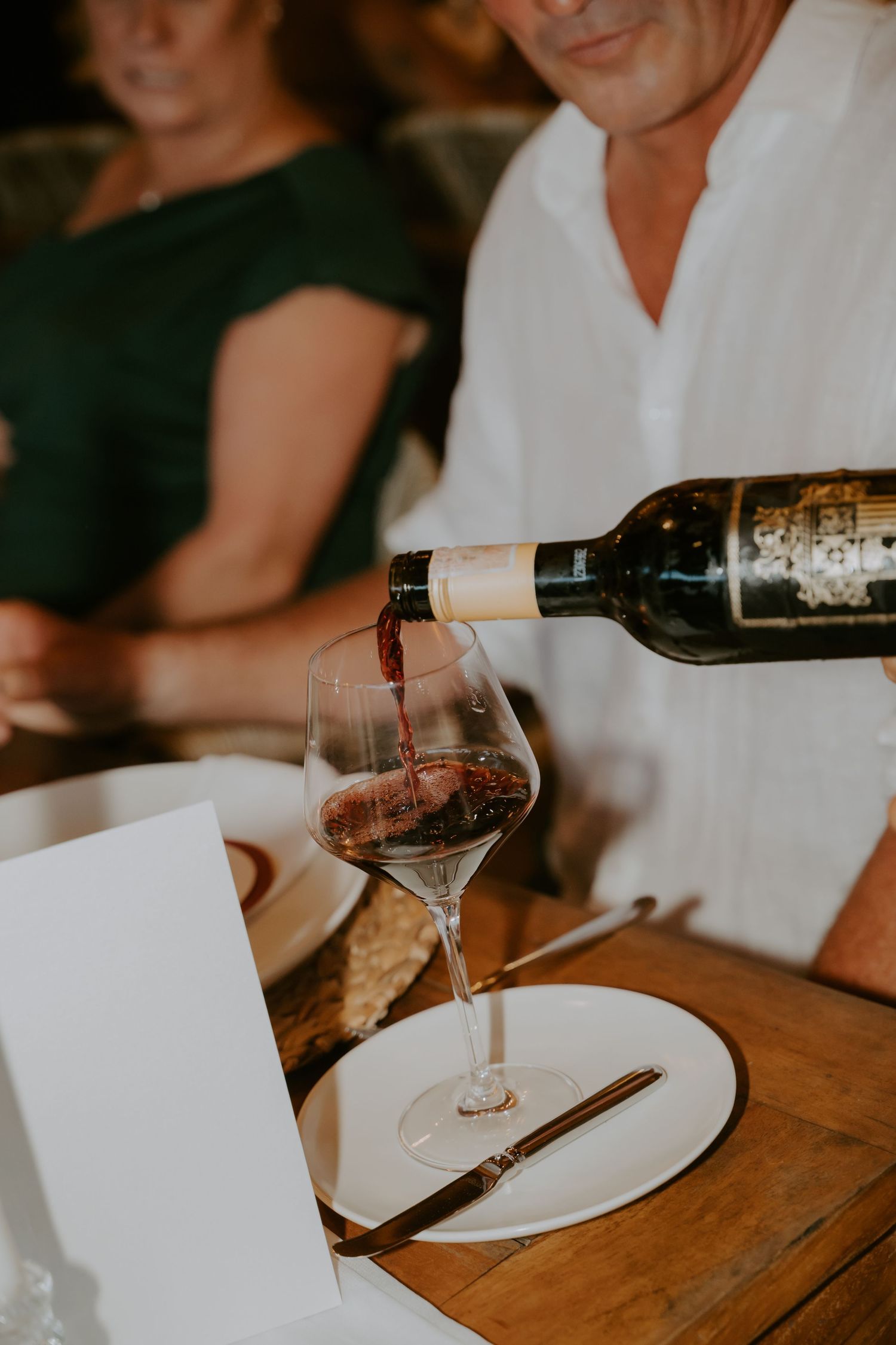 Guest pours red wine into crystal glass at rustic wooden wedding reception table setting.