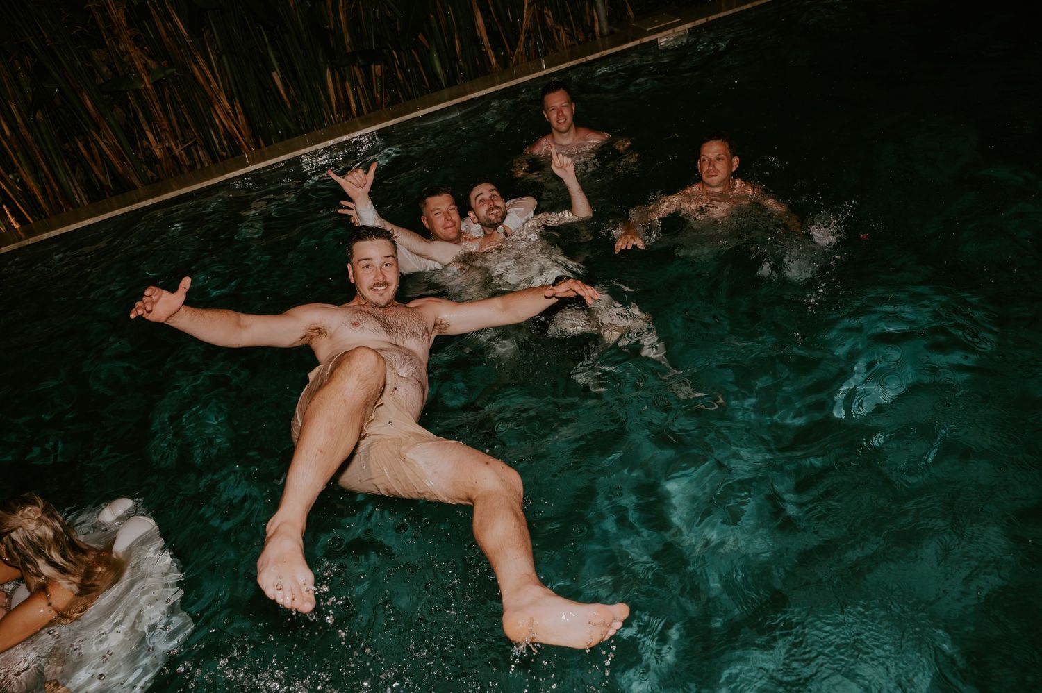 Group of men swimming and celebrating in a pool at a late-night wedding after-party.