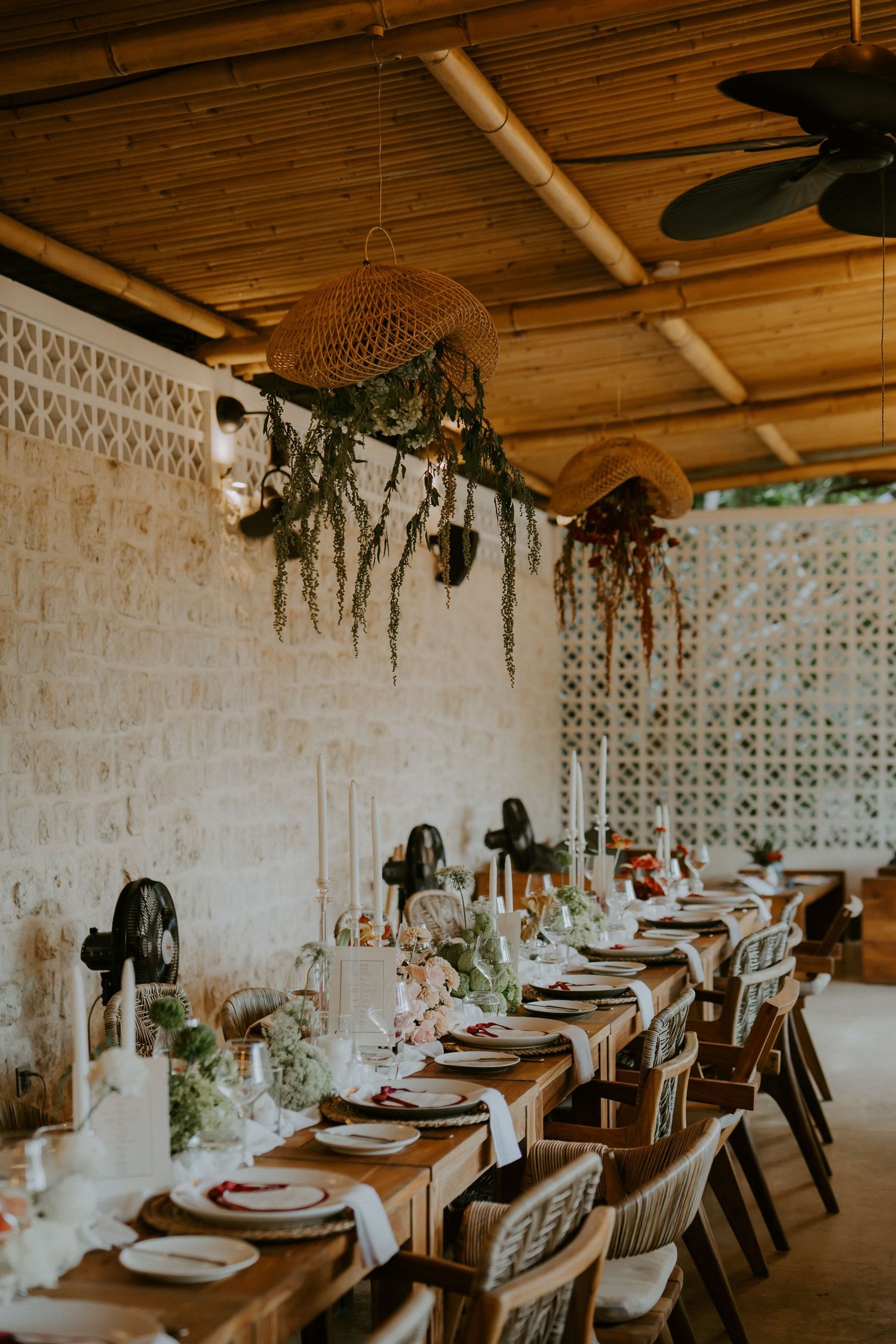 Elegant boho wedding reception with long wooden table, greenery chandelier, and bamboo ceiling in tropical venue.