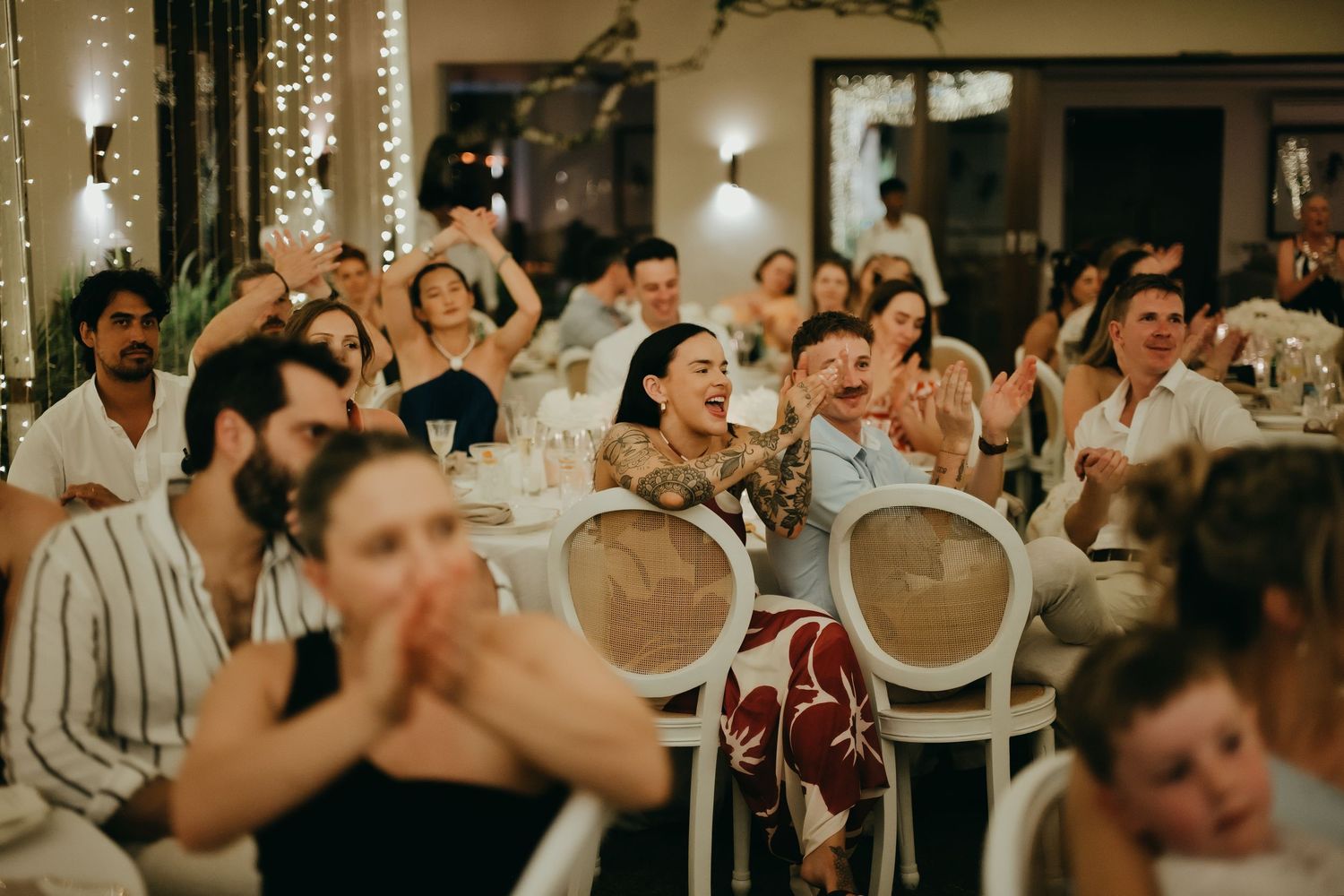 Joyful wedding guests clapping and laughing together during reception speeches at a decorated venue.