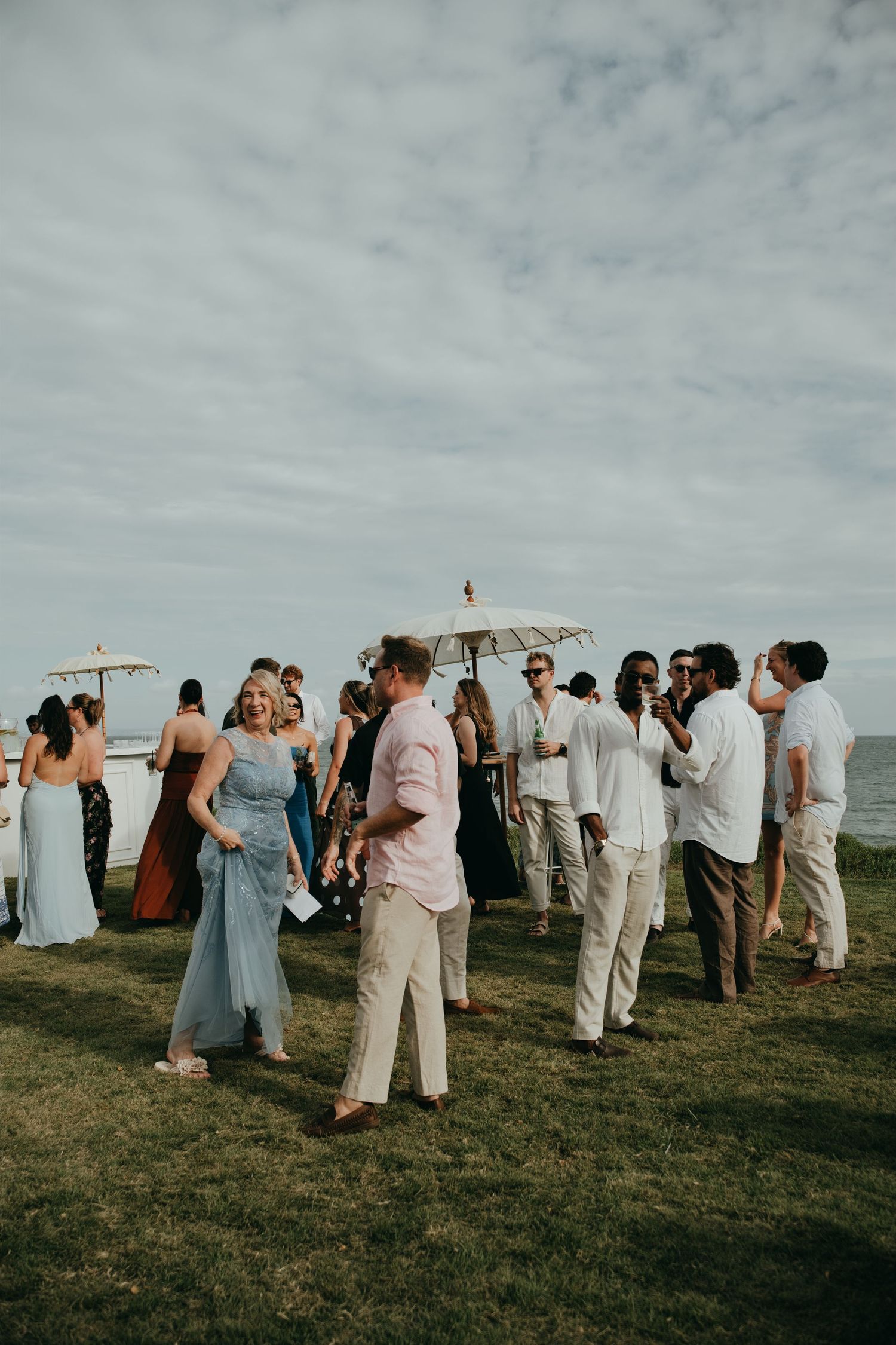 Wedding guests mingling on a clifftop lawn with white parasols and ocean views under a cloudy sky.