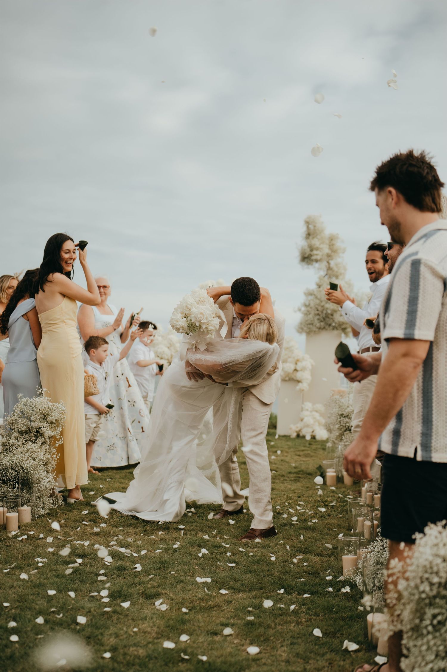 Groom dipping bride during joyful outdoor wedding ceremony exit while guests cheer and toss flower petals.