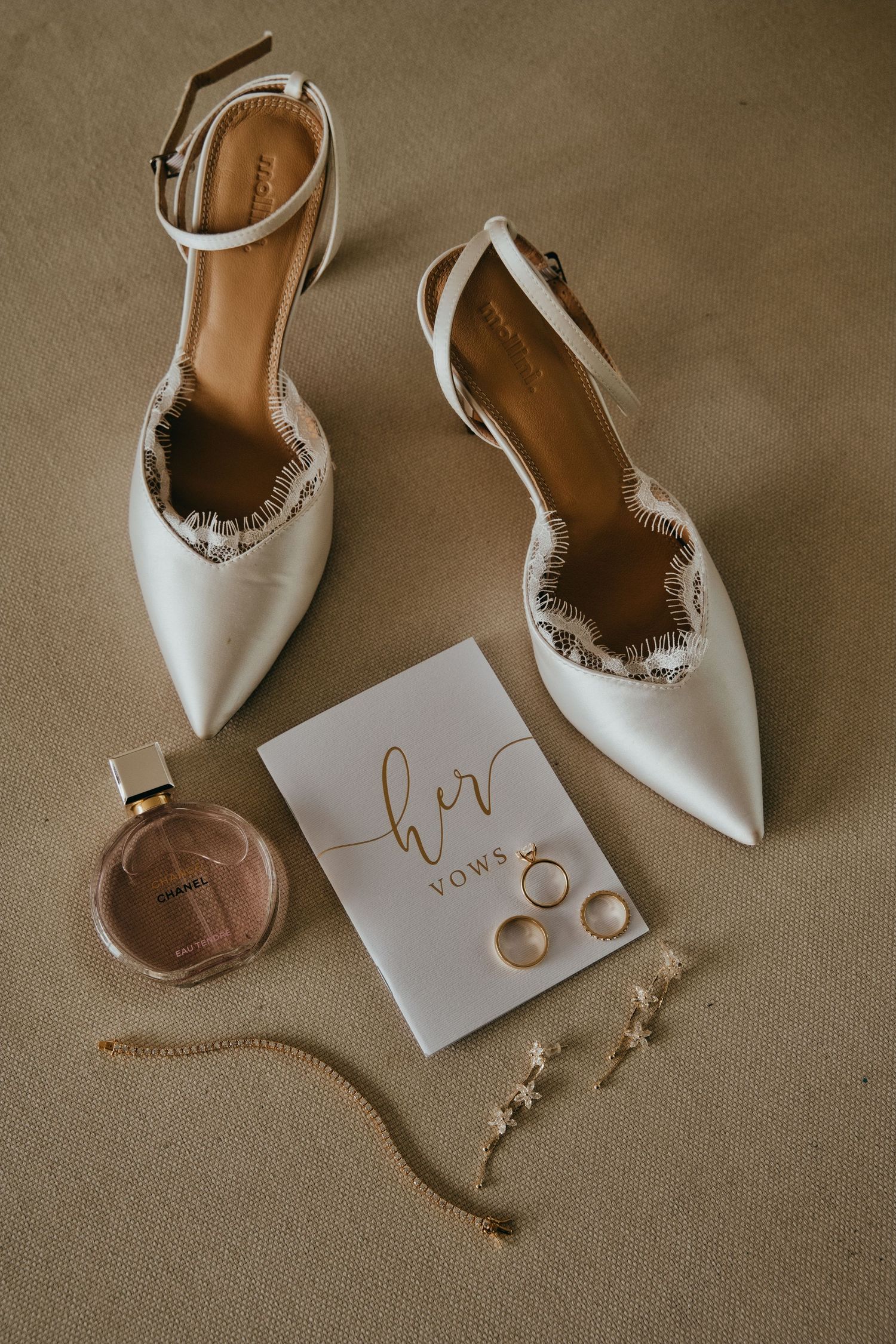 White bridal heels, vow book, wedding rings, pearl earrings, gold bracelet, and Chanel perfume flatlay.