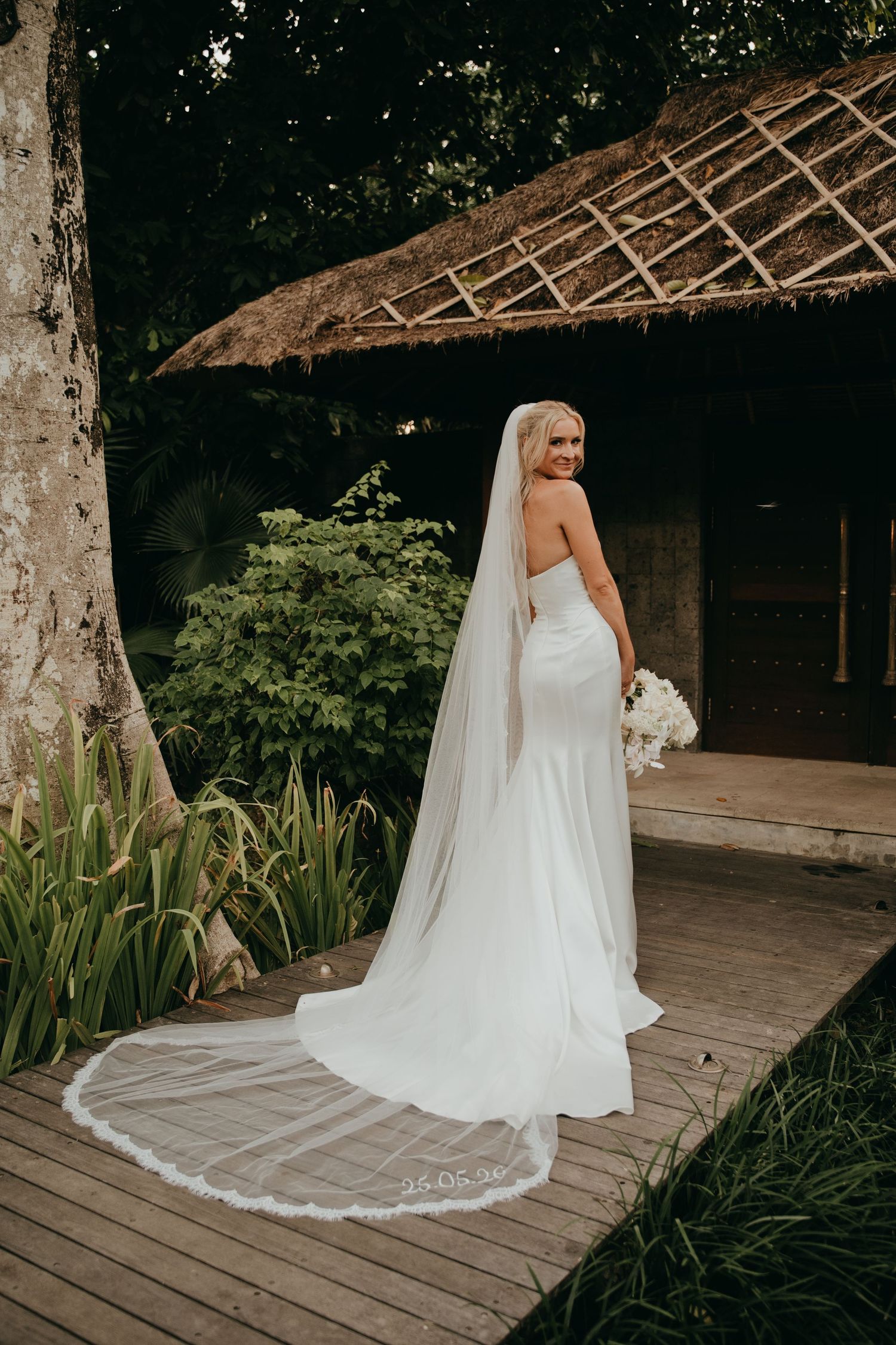 Bride in strapless white gown with long cathedral veil stands on wooden deck surrounded by tropical greenery.