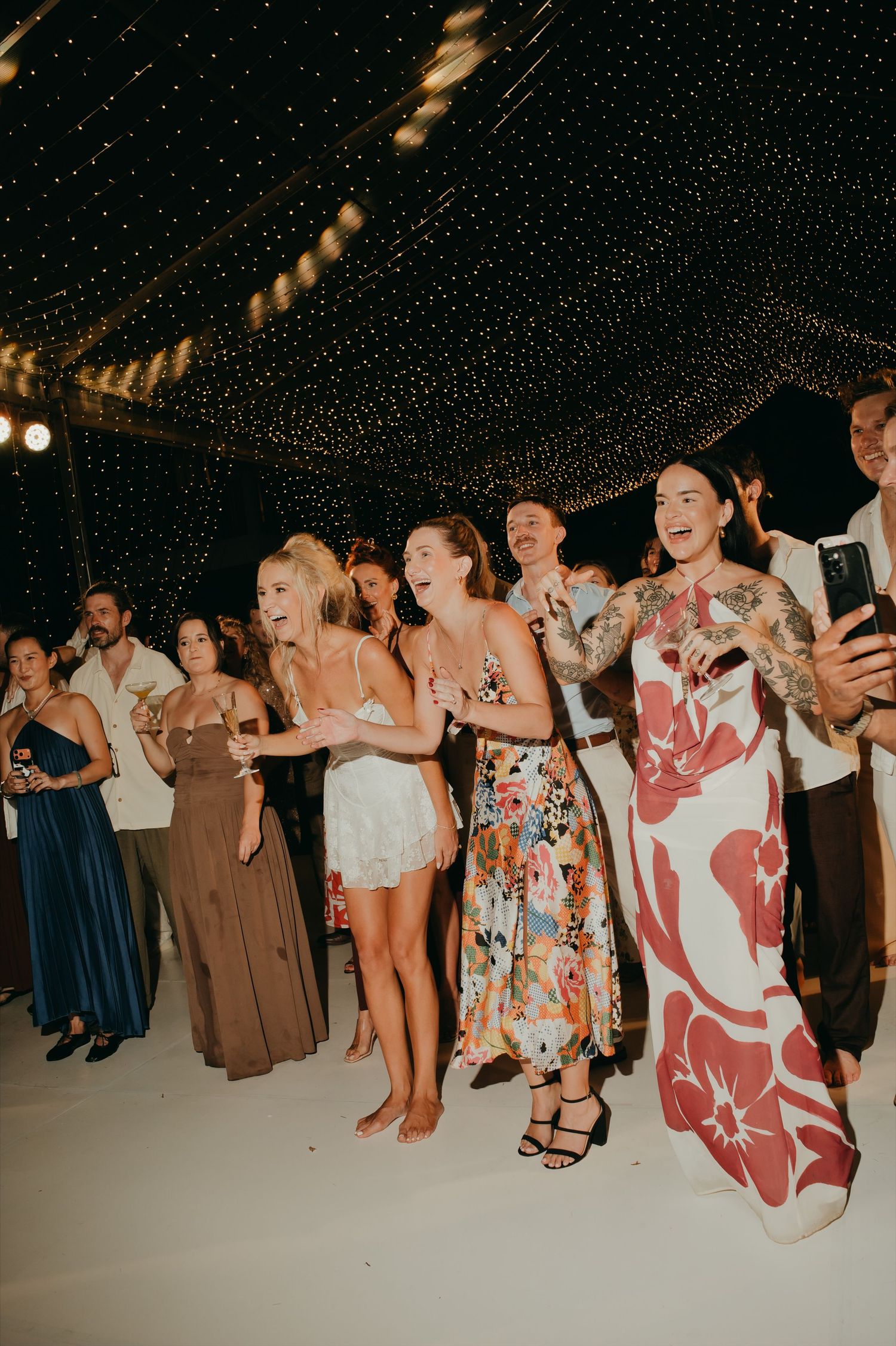 Wedding guests in colorful dresses cheering and taking photos on dance floor under fairy light canopy.