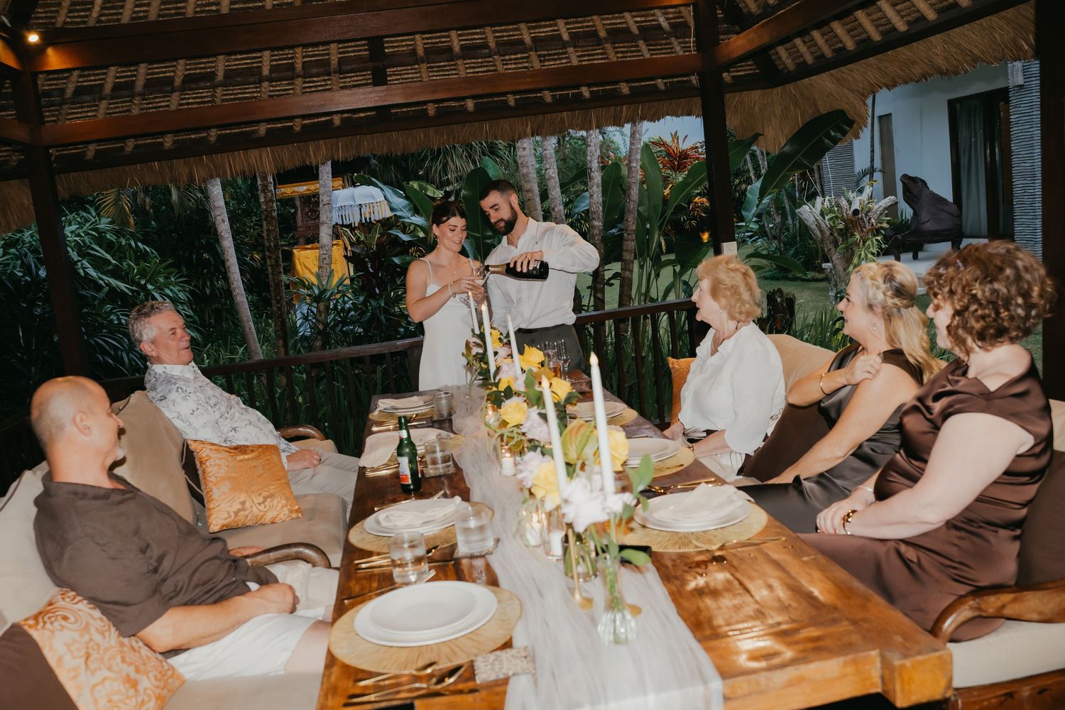 Bride and groom sharing a toast with guests at a warmly lit outdoor wedding reception dinner table.