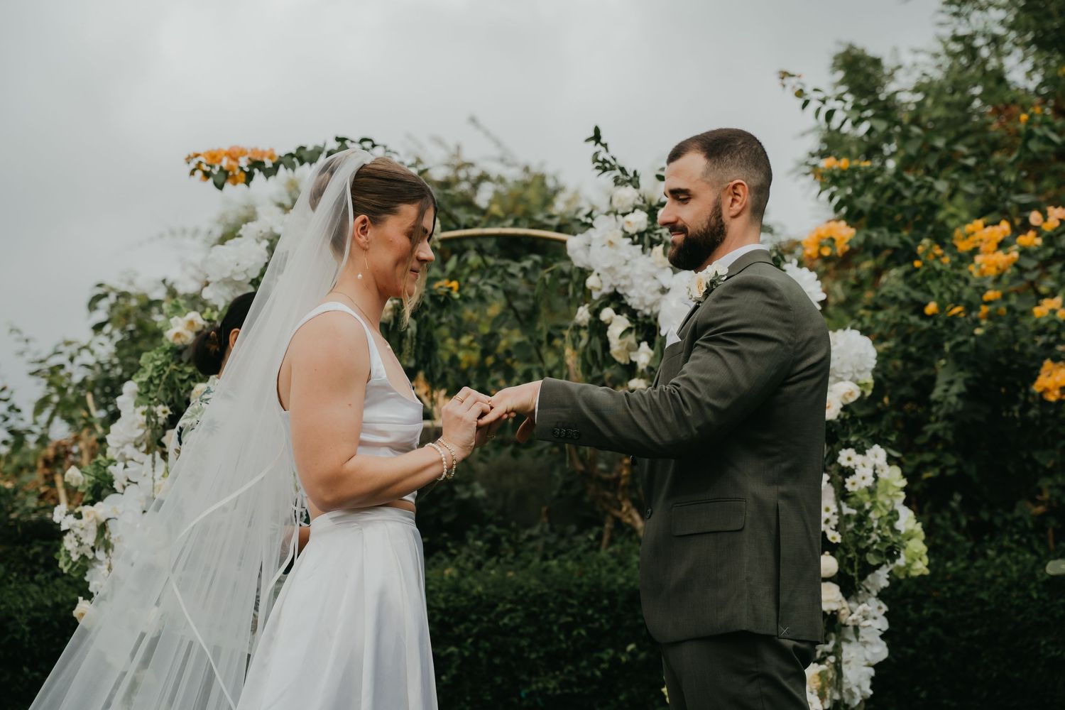 Smiling bride and bearded groom exchange rings under white floral arch during outdoor garden wedding ceremony.