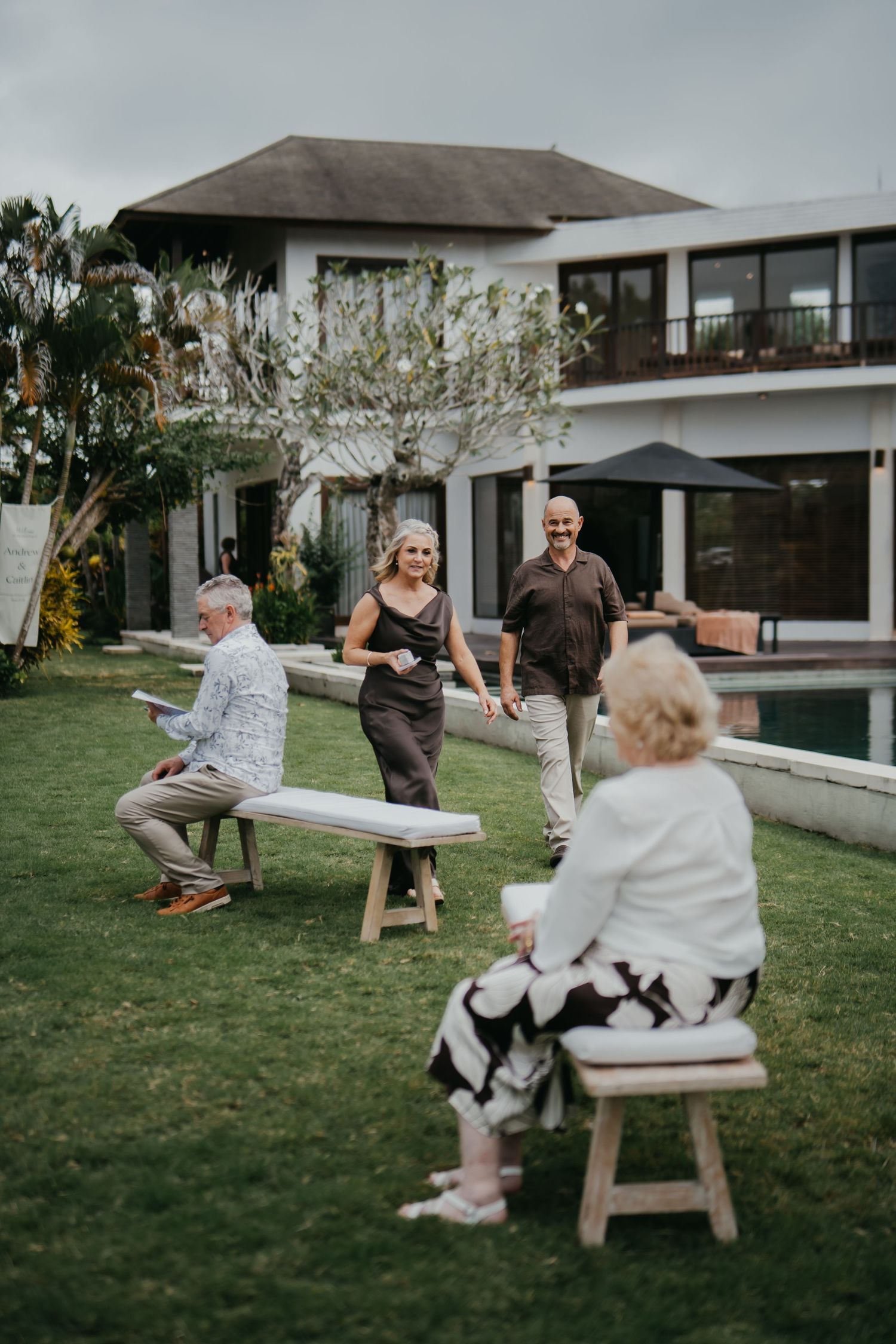 Guests mingling on lush garden lawn of luxury villa with pool during outdoor wedding celebration.