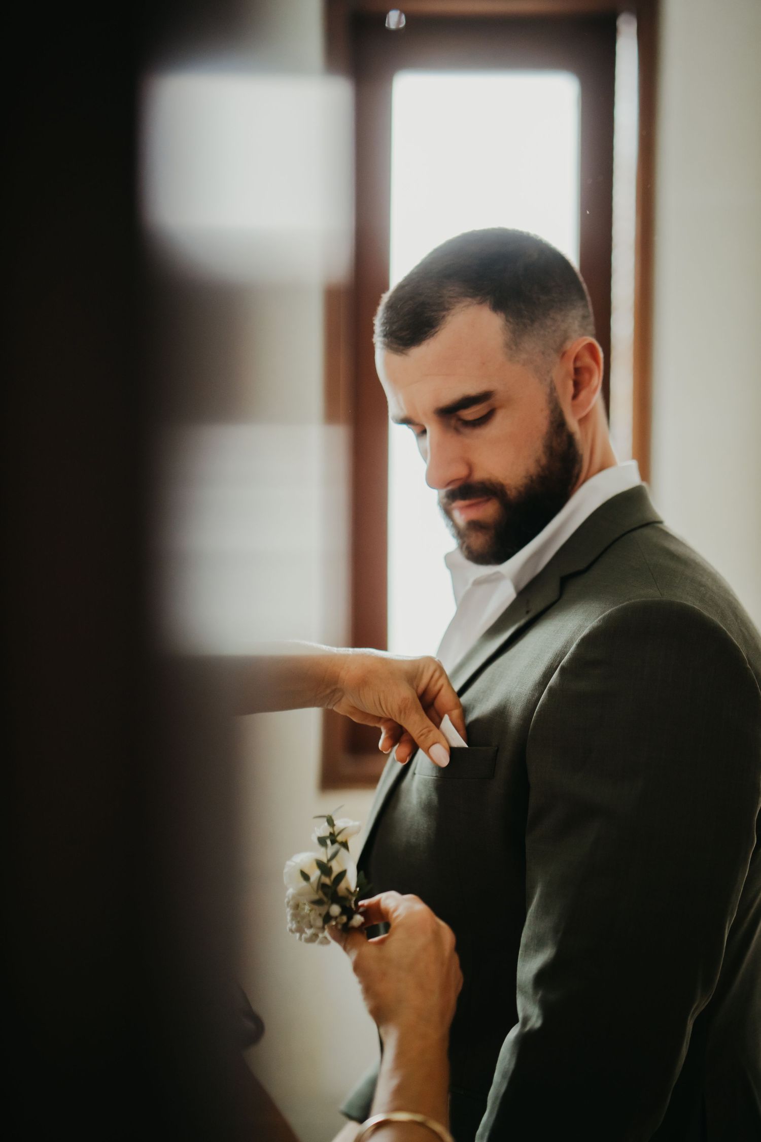 Groom in olive suit getting boutonniere pinned before wedding ceremony.